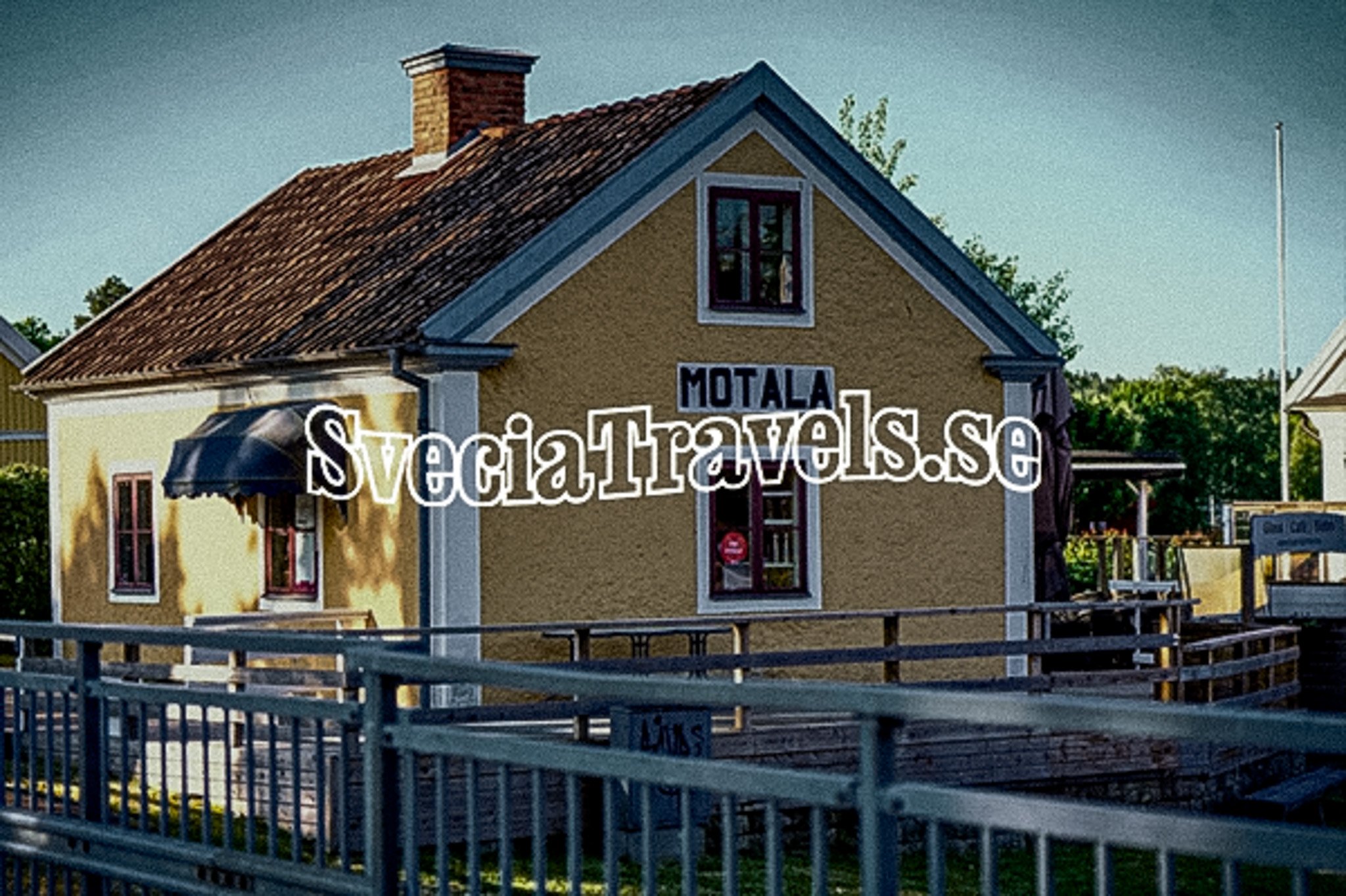 A yellow building with a brown tiled roof, "MOTALA" sign, and a fence in the foreground.