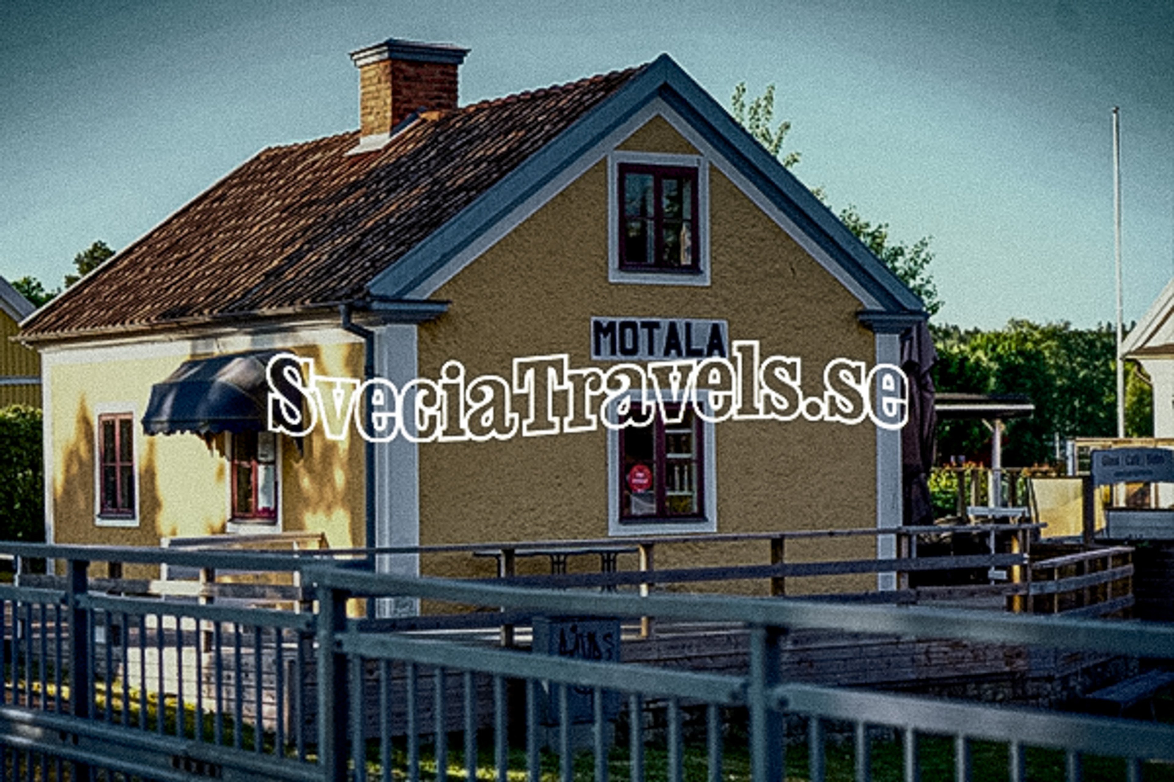 A yellow building with a brown tiled roof, "MOTALA" sign, and a fence in the foreground.