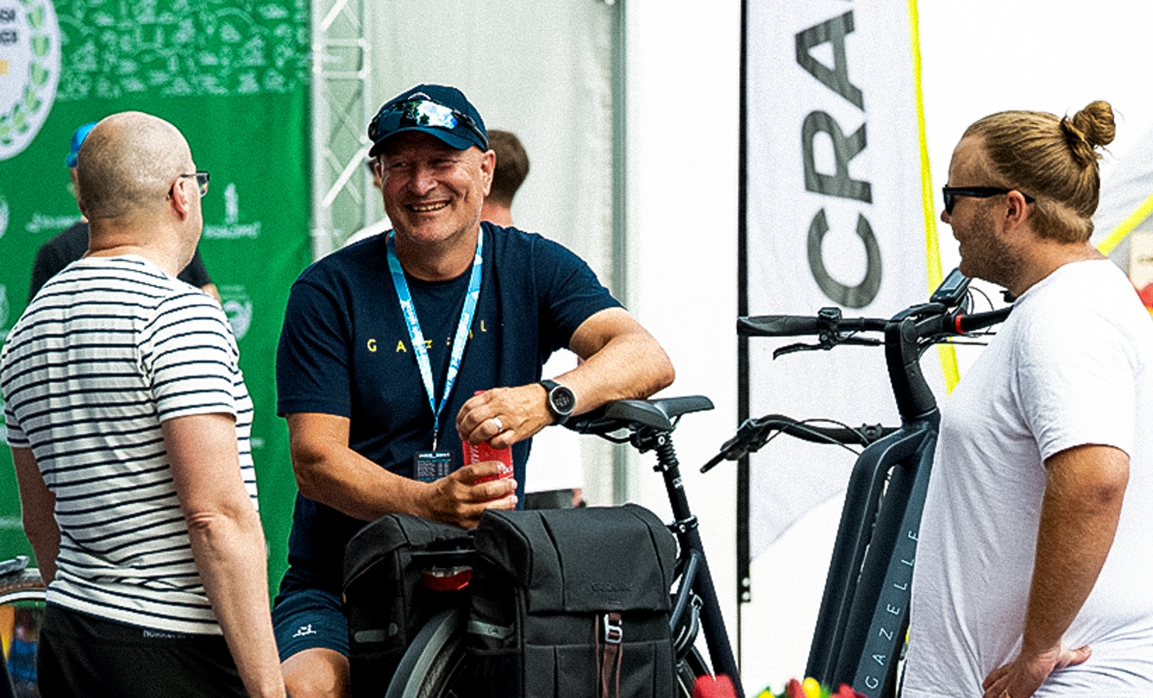 Three men talking and smiling outdoors next to bicycles; one man holds a red can.