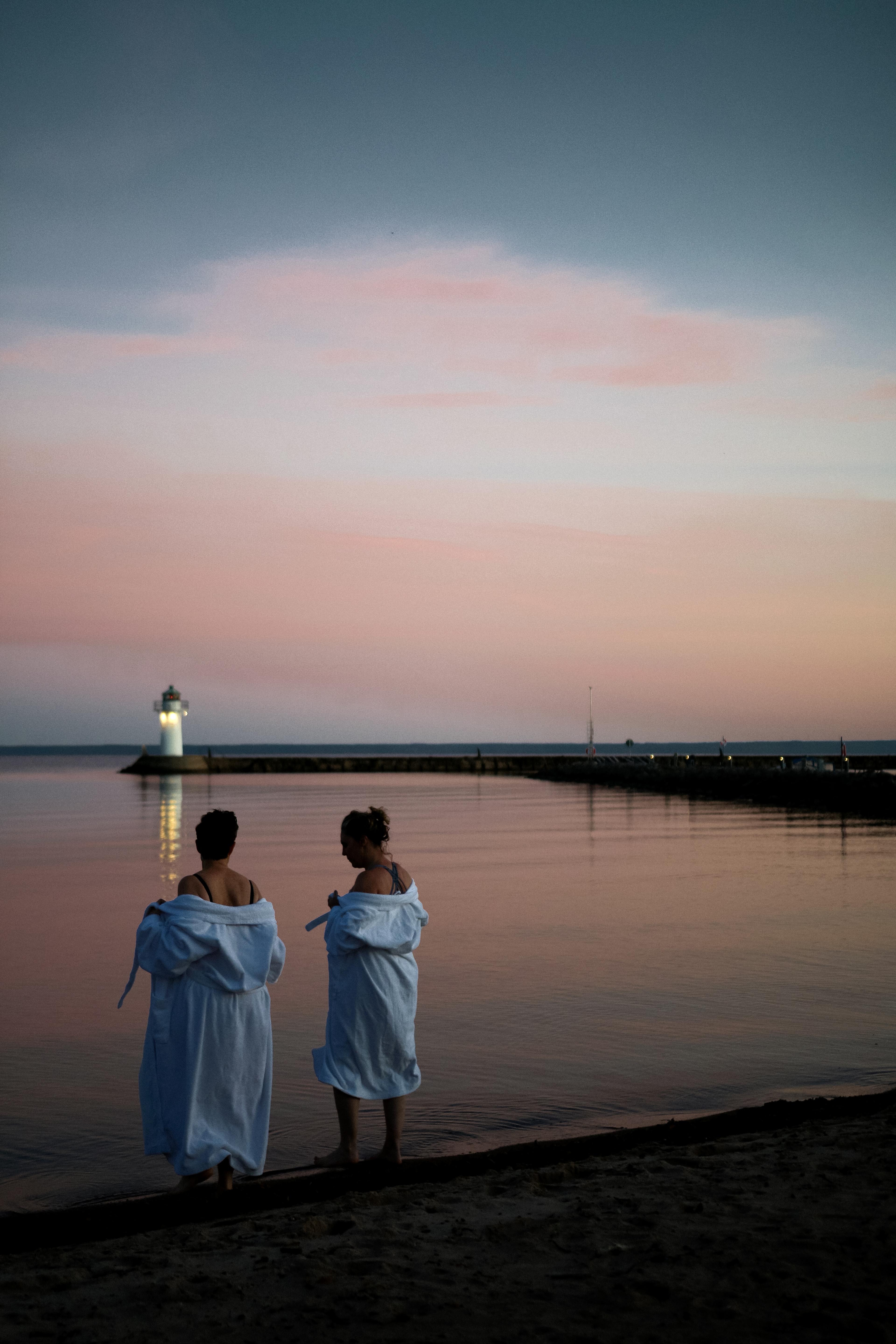Two women in white bathrobes stand by the water's edge at sunset, with a lighthouse and pier in the distance.