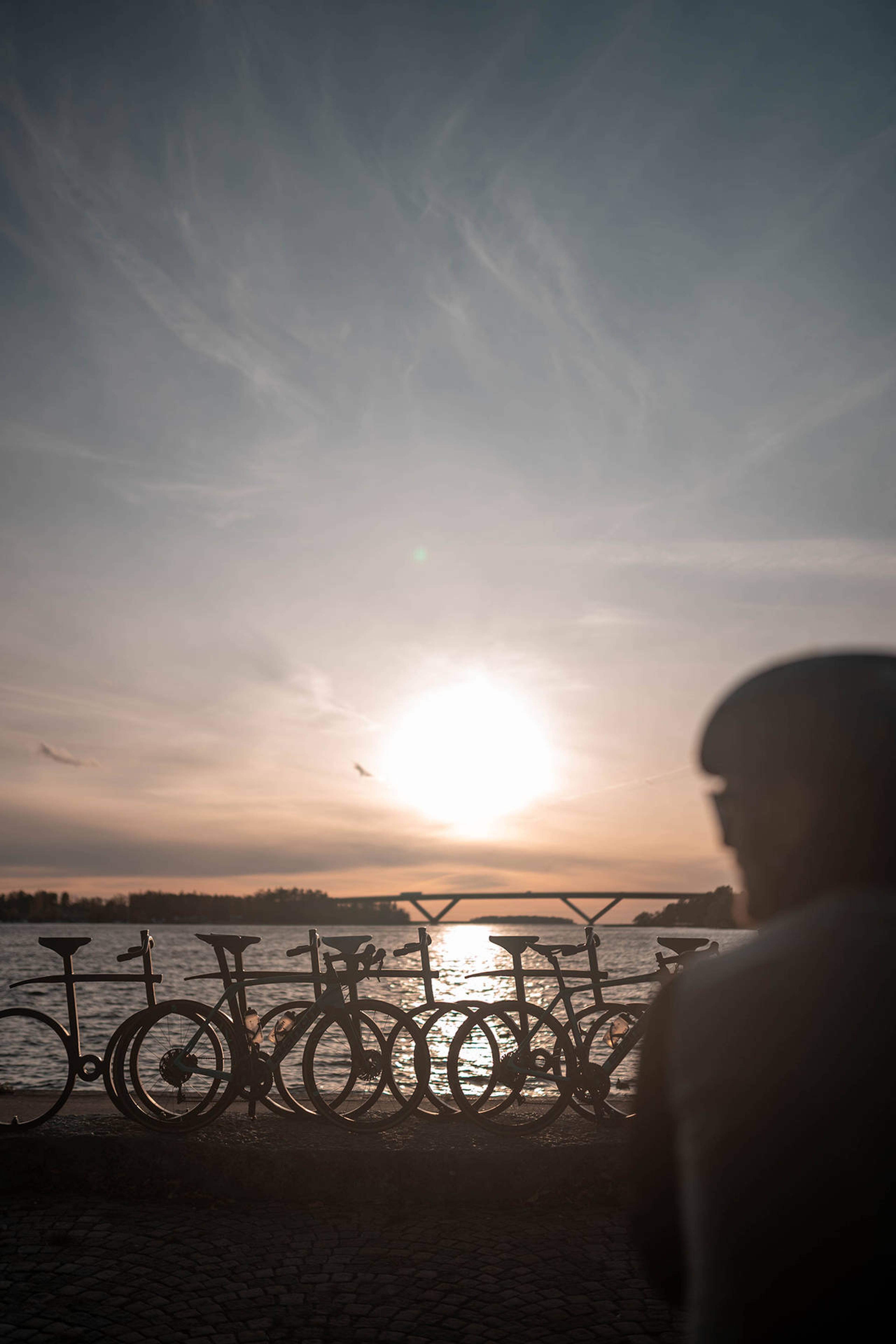 Silhouettes of bicycles and a person in front of a bright sunset over water with a distant bridge.