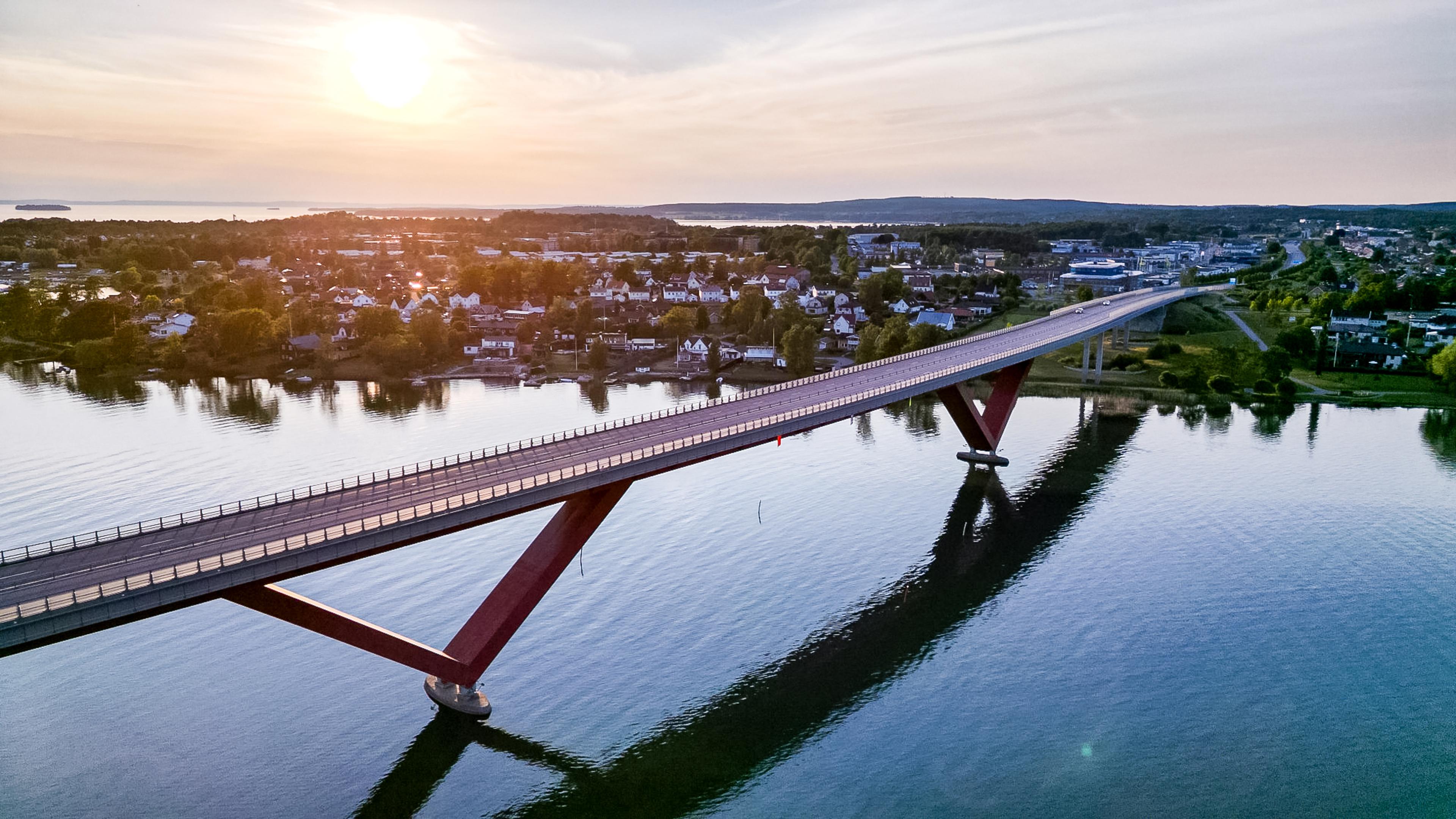 A modern bridge with a red V-shaped support spans water, connecting towns, under a sunset sky.