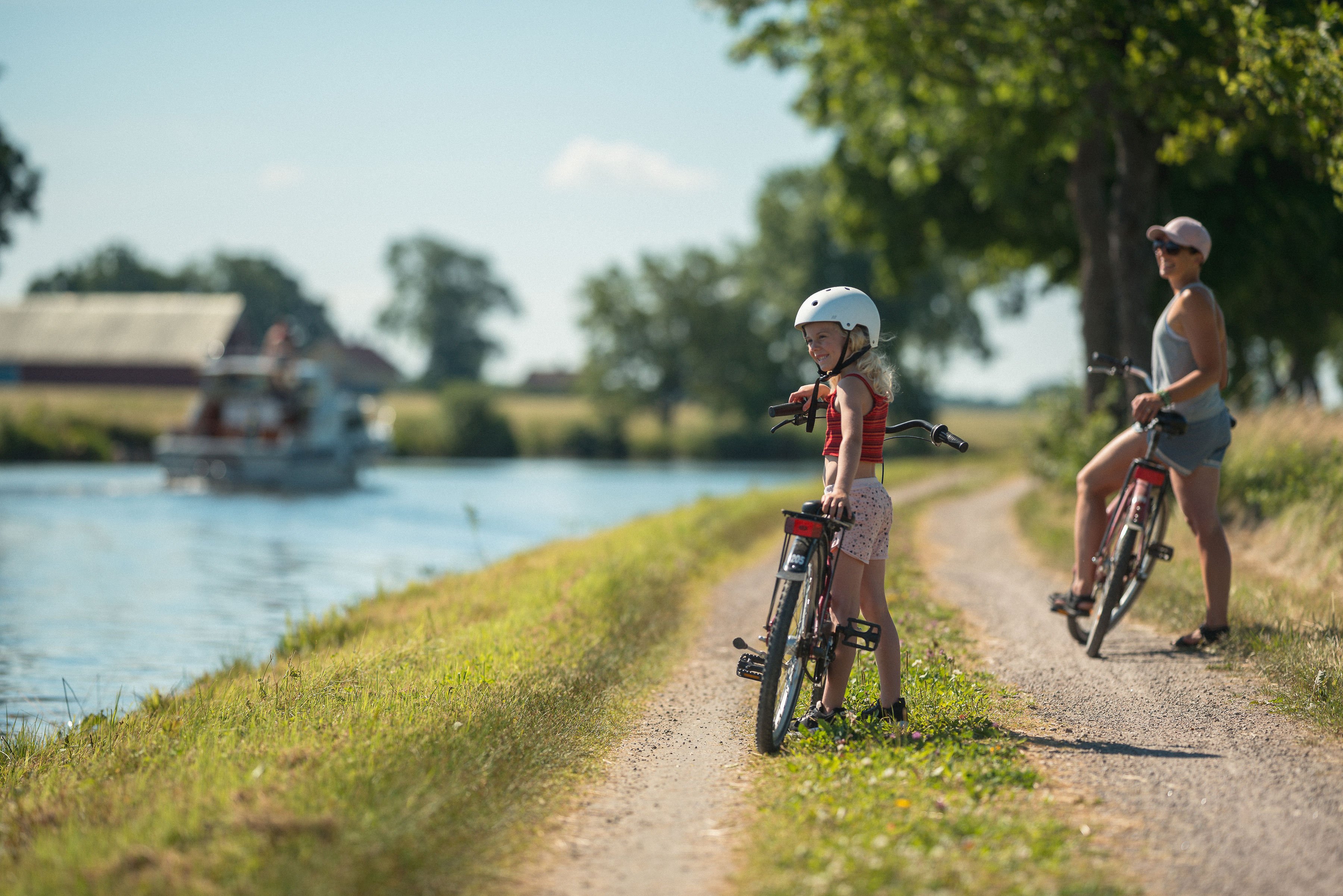 A young girl and an adult woman with bikes on a dirt path next to a canal with a boat.