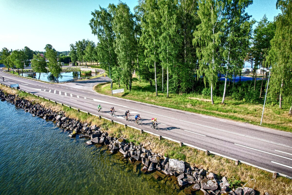 A group of cyclists rides along a road next to a rocky lake shore lined with tall trees on a clear day.