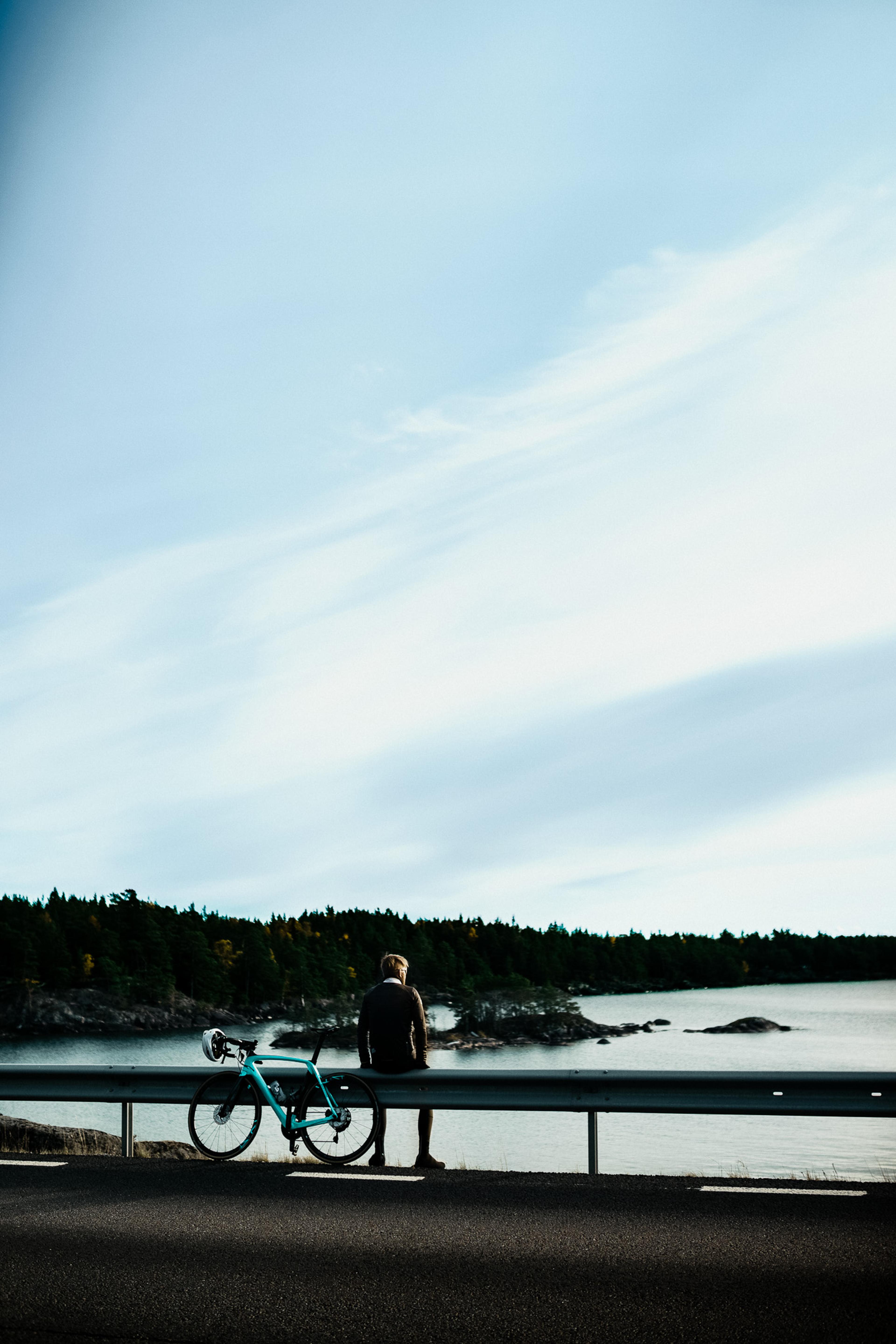 A person sits on a guardrail with a teal bicycle nearby, overlooking a lake and forest under a wide sky.