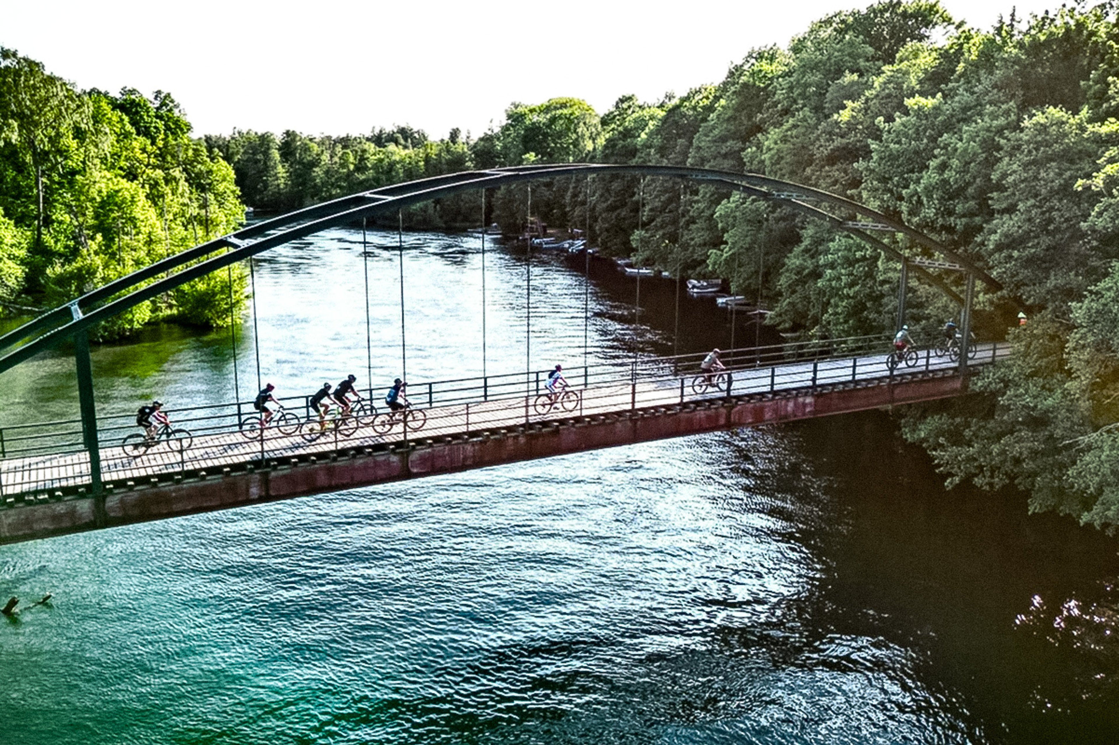 Seven cyclists ride across an arch bridge over a river with lush trees.