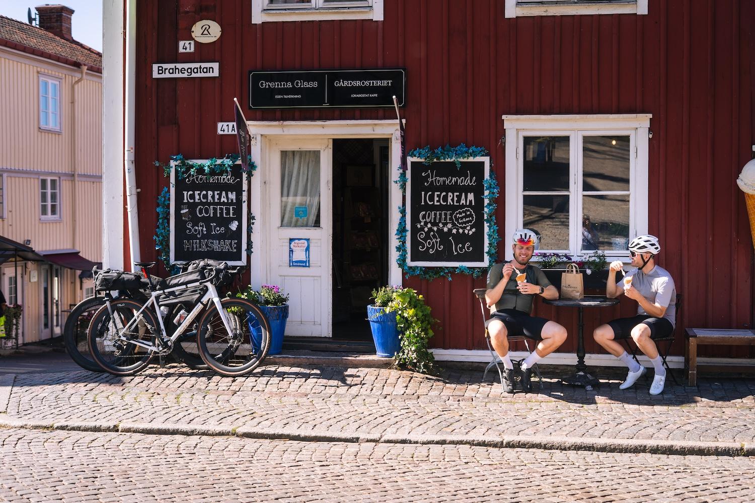Two cyclists in helmets and gear enjoy ice cream outside a red ice cream shop, with their bikes parked nearby on a cobblestone street.