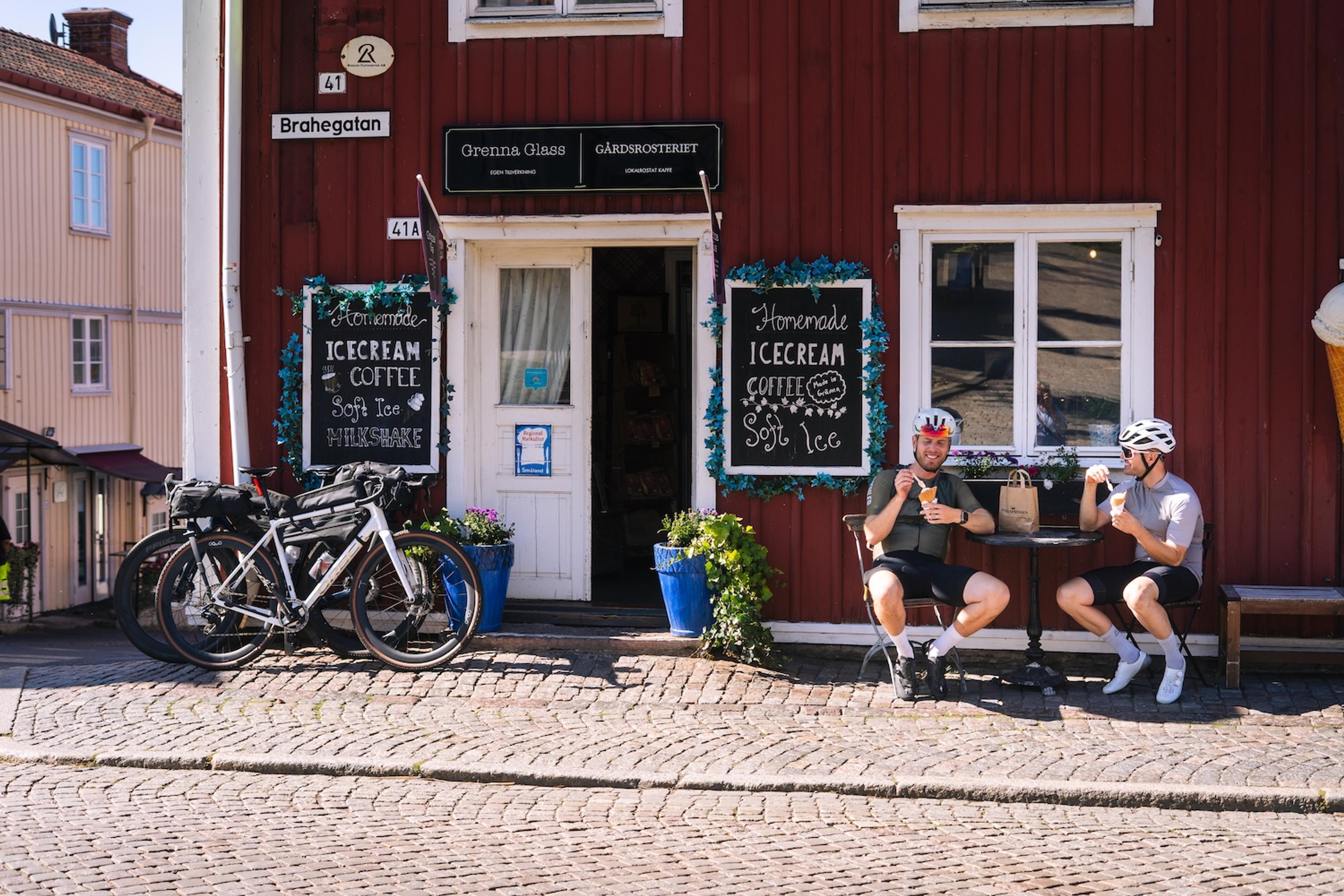 Two cyclists in helmets and gear enjoy ice cream outside a red ice cream shop, with their bikes parked nearby on a cobblestone street.