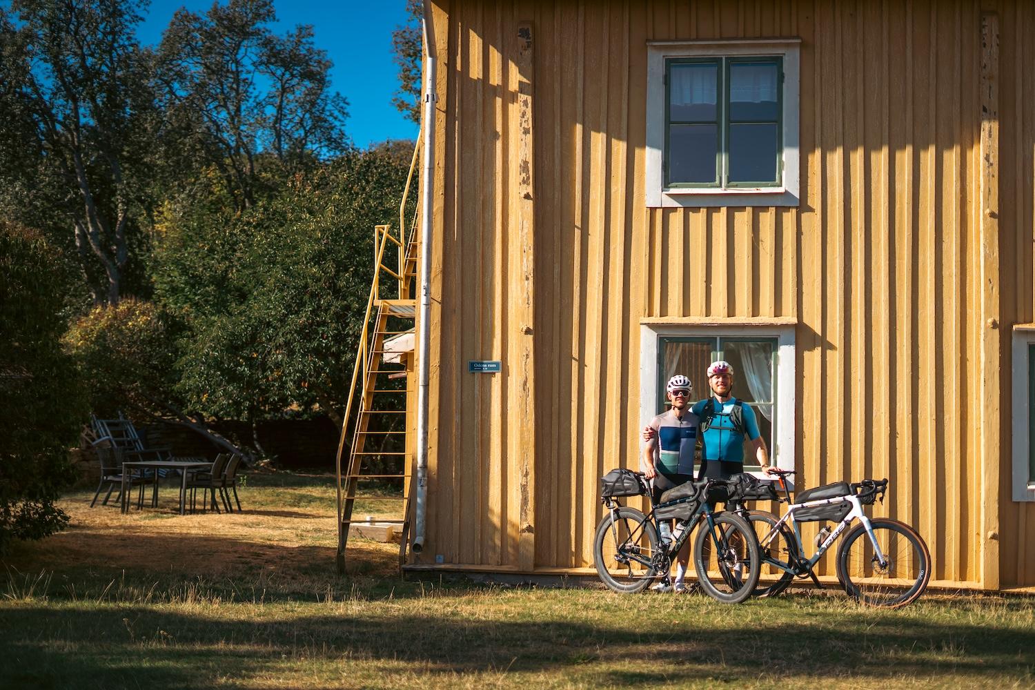 Two cyclists with bikepacking bikes stand in front of a yellow house.