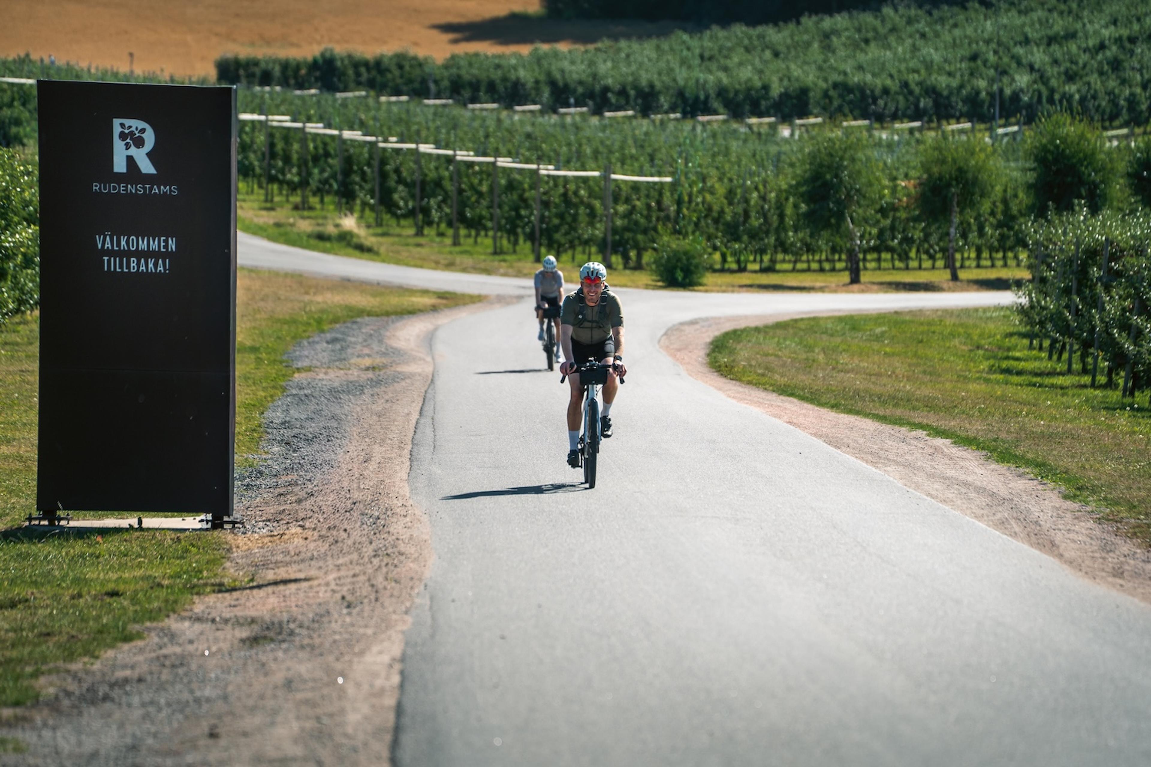 Two cyclists ride on a winding road through orchards, past a welcome sign for Rudenstams.