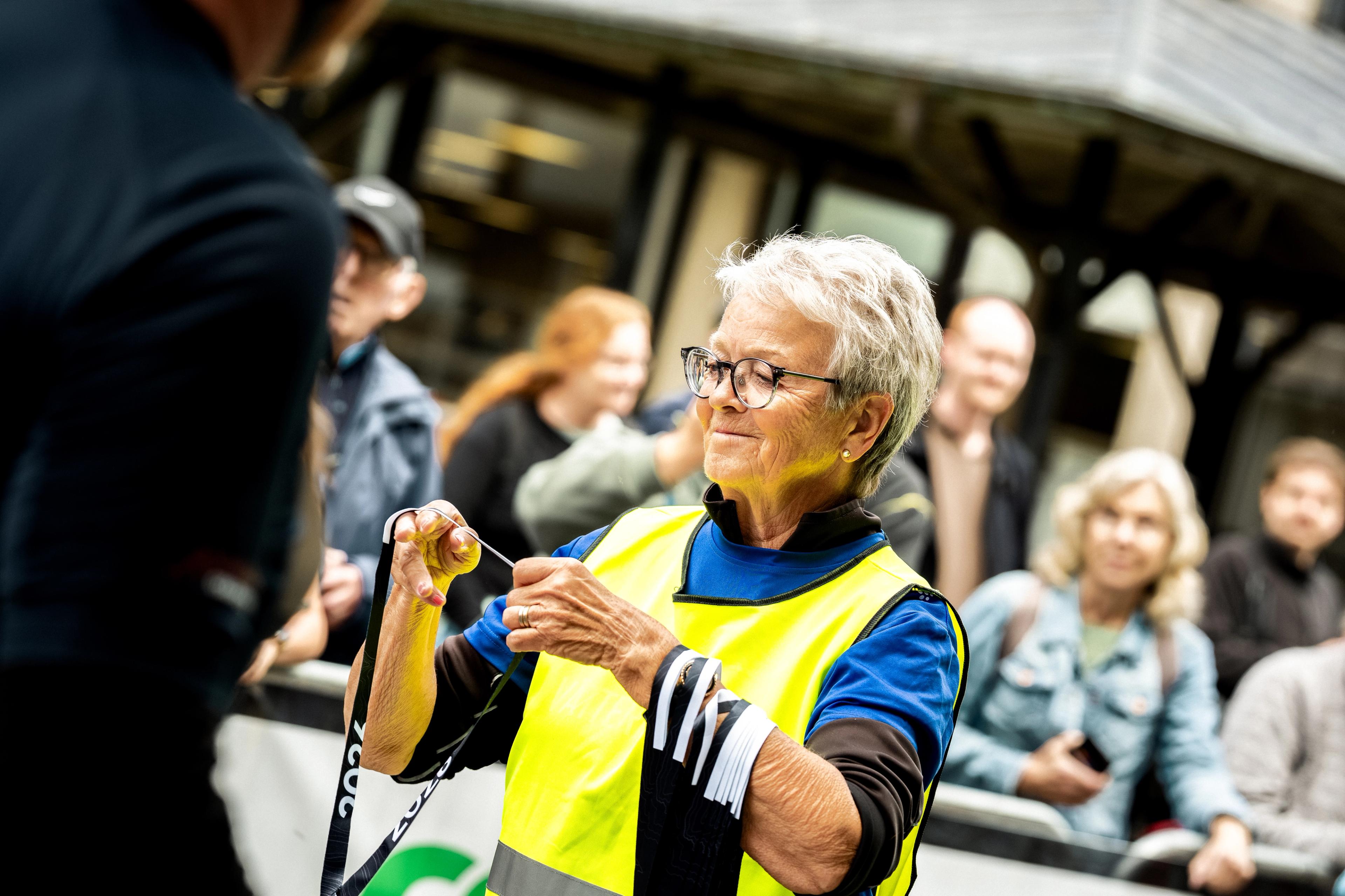 An older woman in a yellow safety vest and glasses sorts black and white lanyards at an event.An older woman in a yellow safety vest and glasses holds black and white striped ribbons.