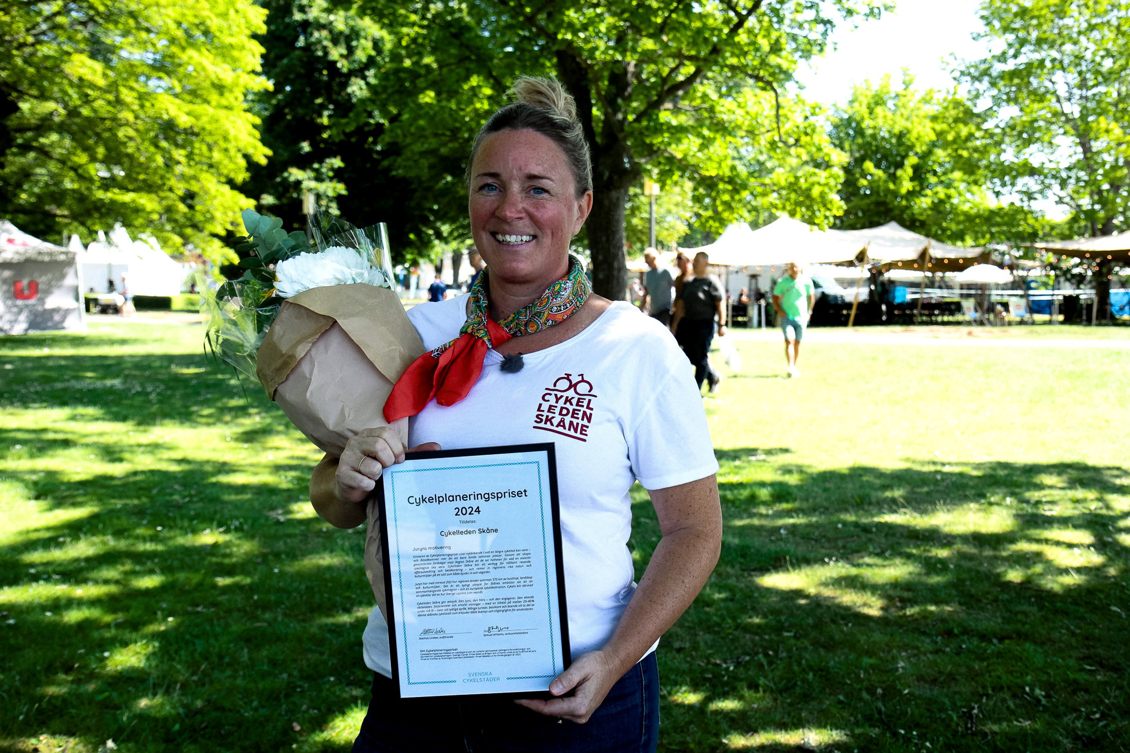 Smiling woman holds a framed "Bicycle Planning Award 2024" certificate and a bouquet of flowers in an outdoor park.