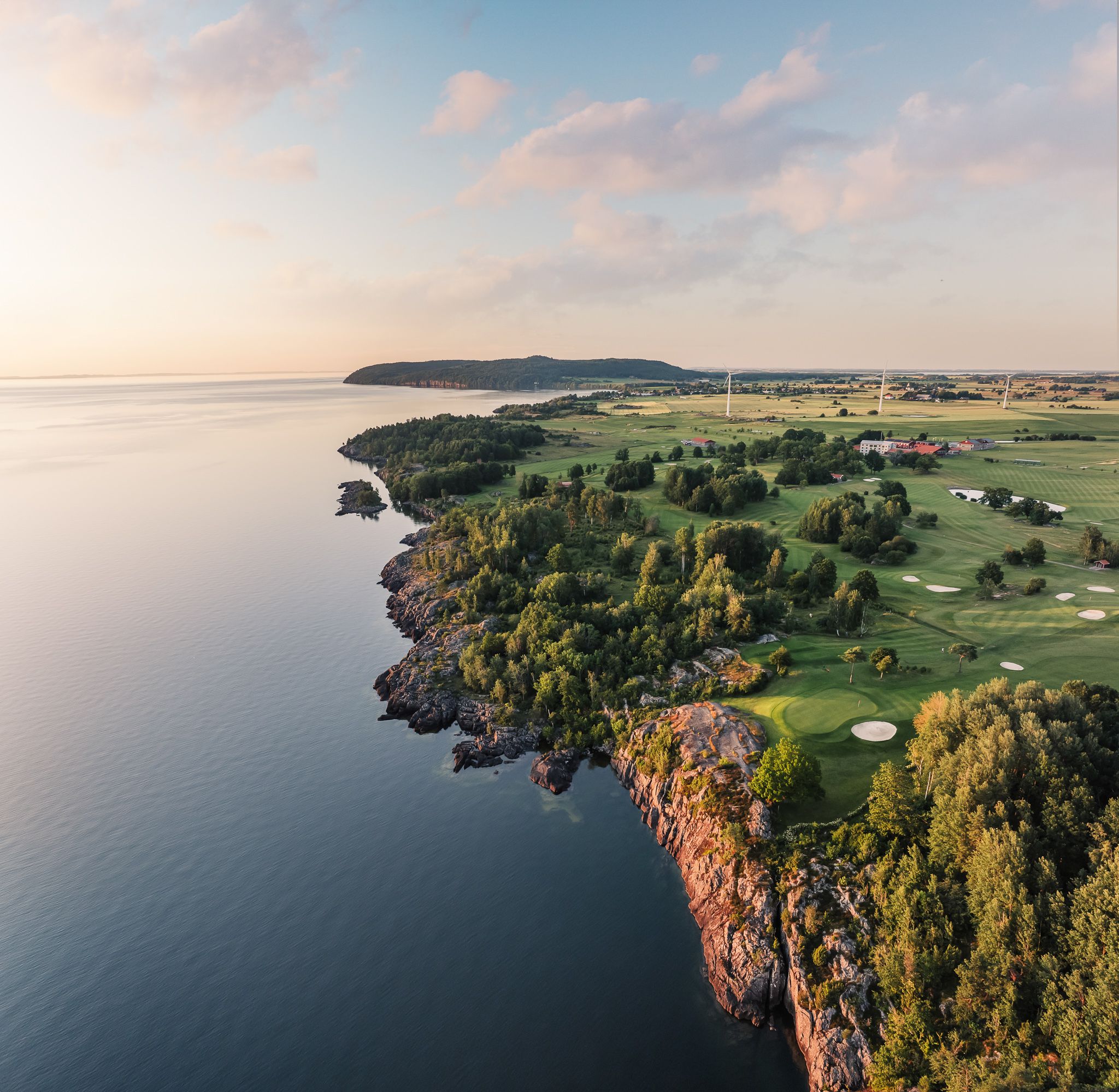 Aerial view of a coastal golf course and rocky shore next to calm water, under a soft sky.