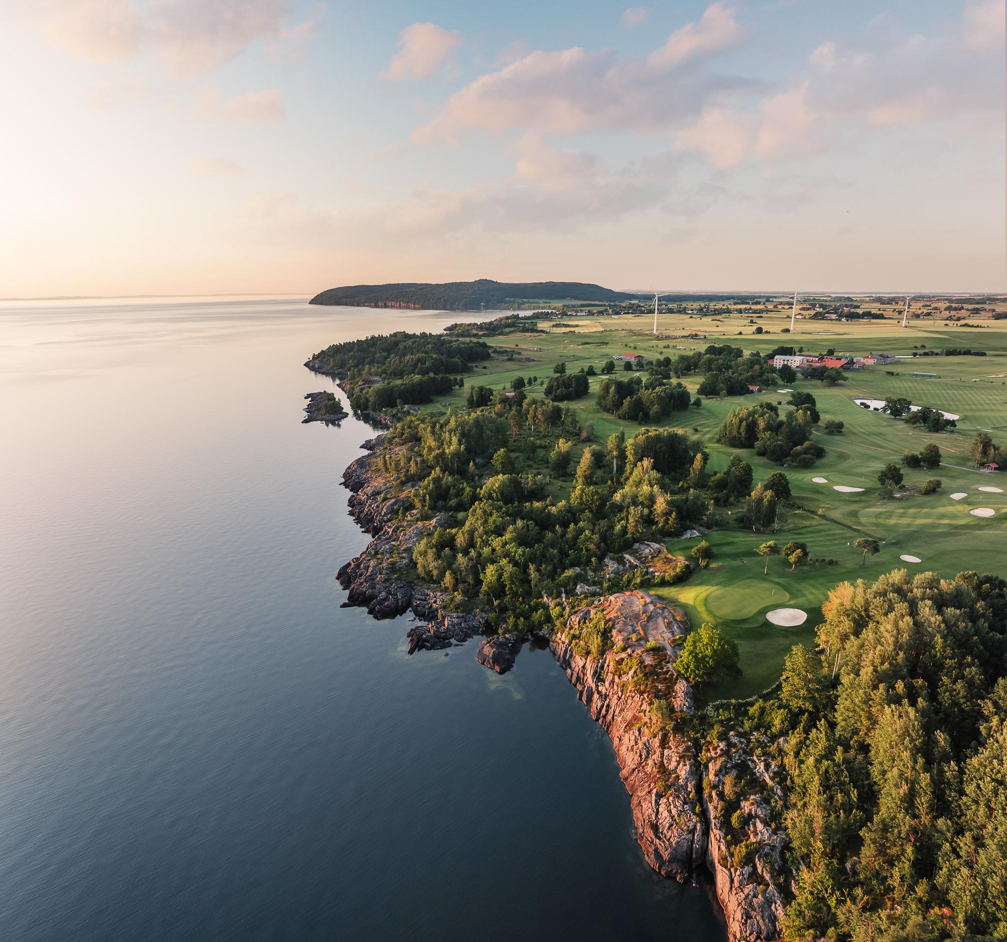 Aerial view of a coastal golf course and rocky shore next to calm water, under a soft sky.