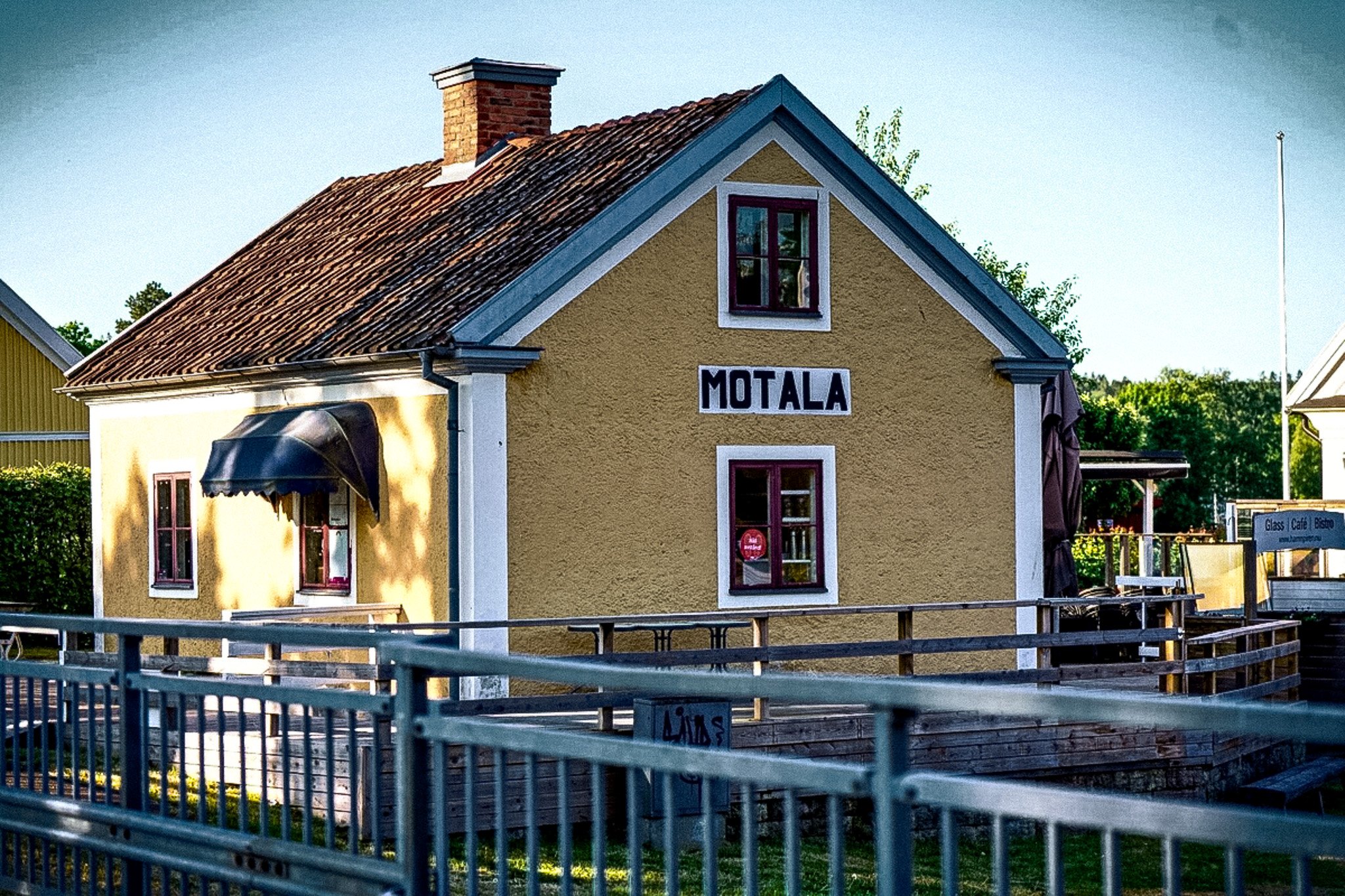 A yellow building with a red tile roof, "MOTALA" sign, and dark awning, next to a wooden dock with a metal railing.