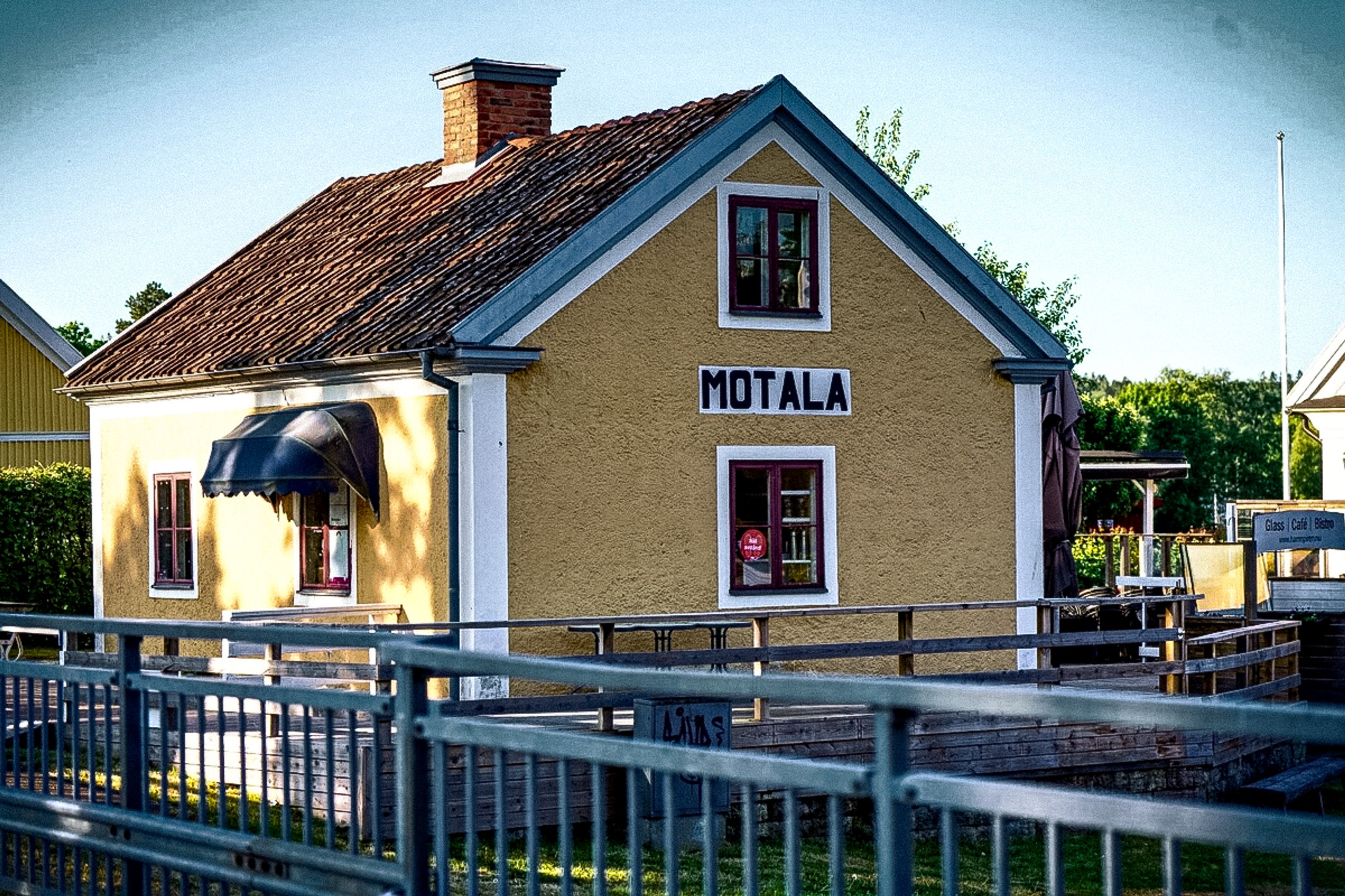 A yellow building with a red tile roof, "MOTALA" sign, and dark awning, next to a wooden dock with a metal railing.