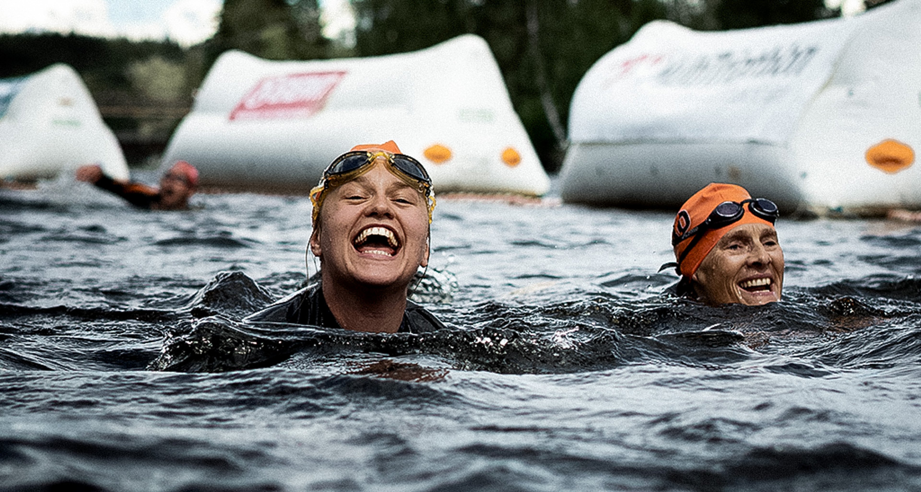 Two women in orange swim caps and goggles smiling and laughing in open water, with event buoys behind them.
