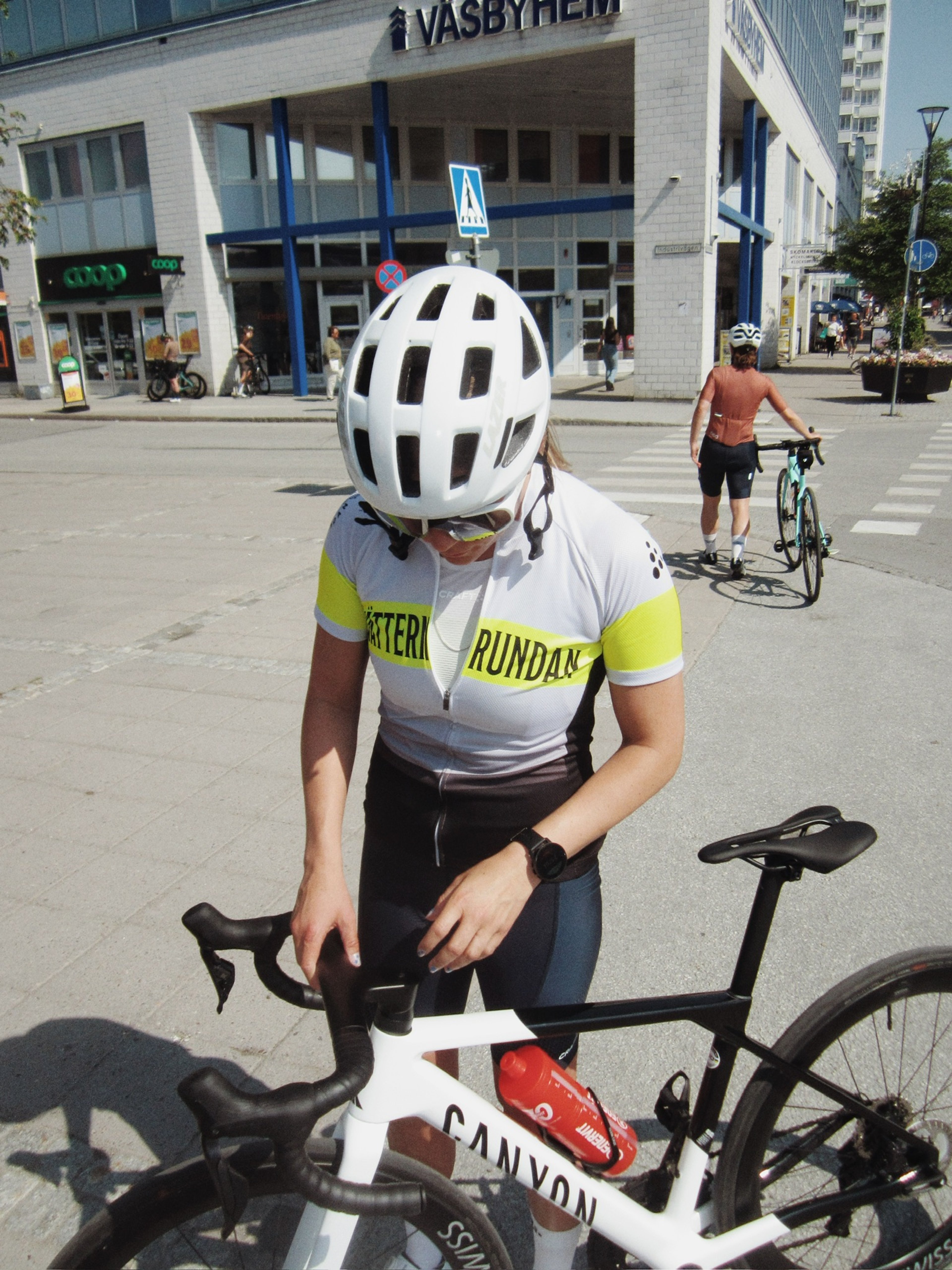 A cyclist in a white helmet and grey and yellow jersey stands next to a white Canyon road bike in an urban setting.