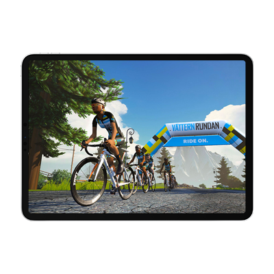 A tablet displaying a cycling simulation game with avatars on a road passing under a "Vätternrundan Ride On" archway.