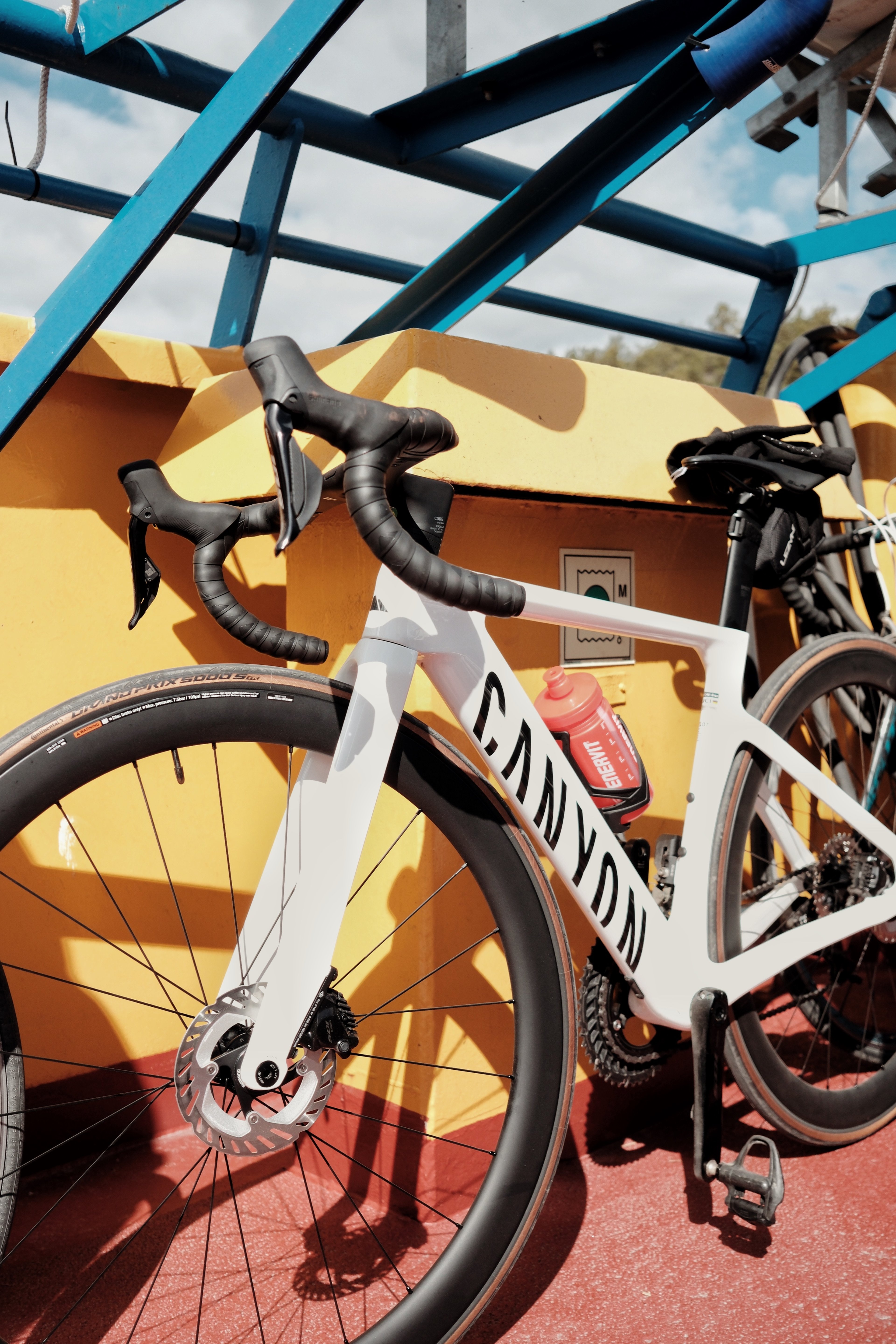 A white Canyon road bike with a red water bottle leans against a yellow wall, with blue beams overhead.