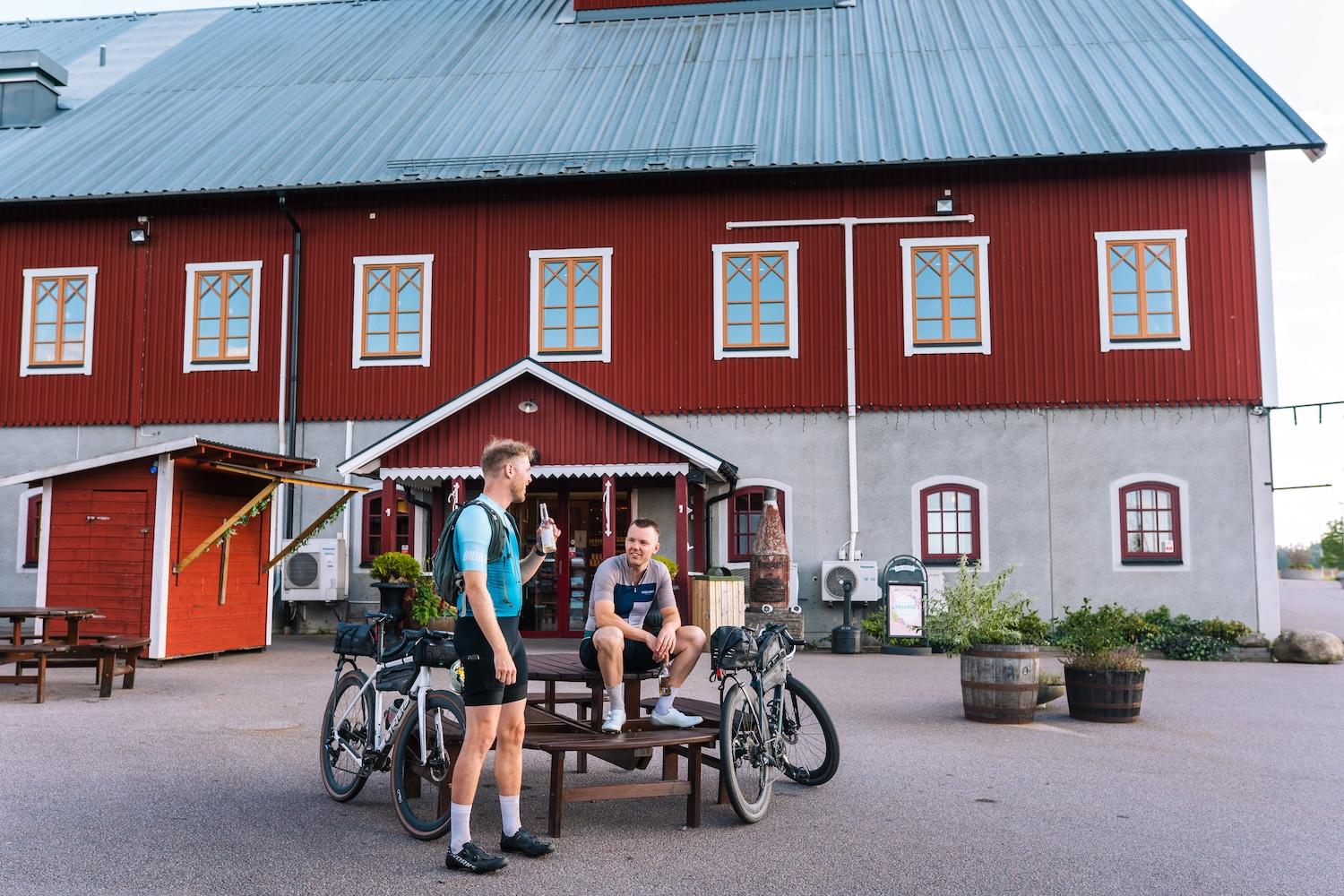 Two cyclists with bikes resting on a patio in front of a red building. One stands with a drink, the other sits.