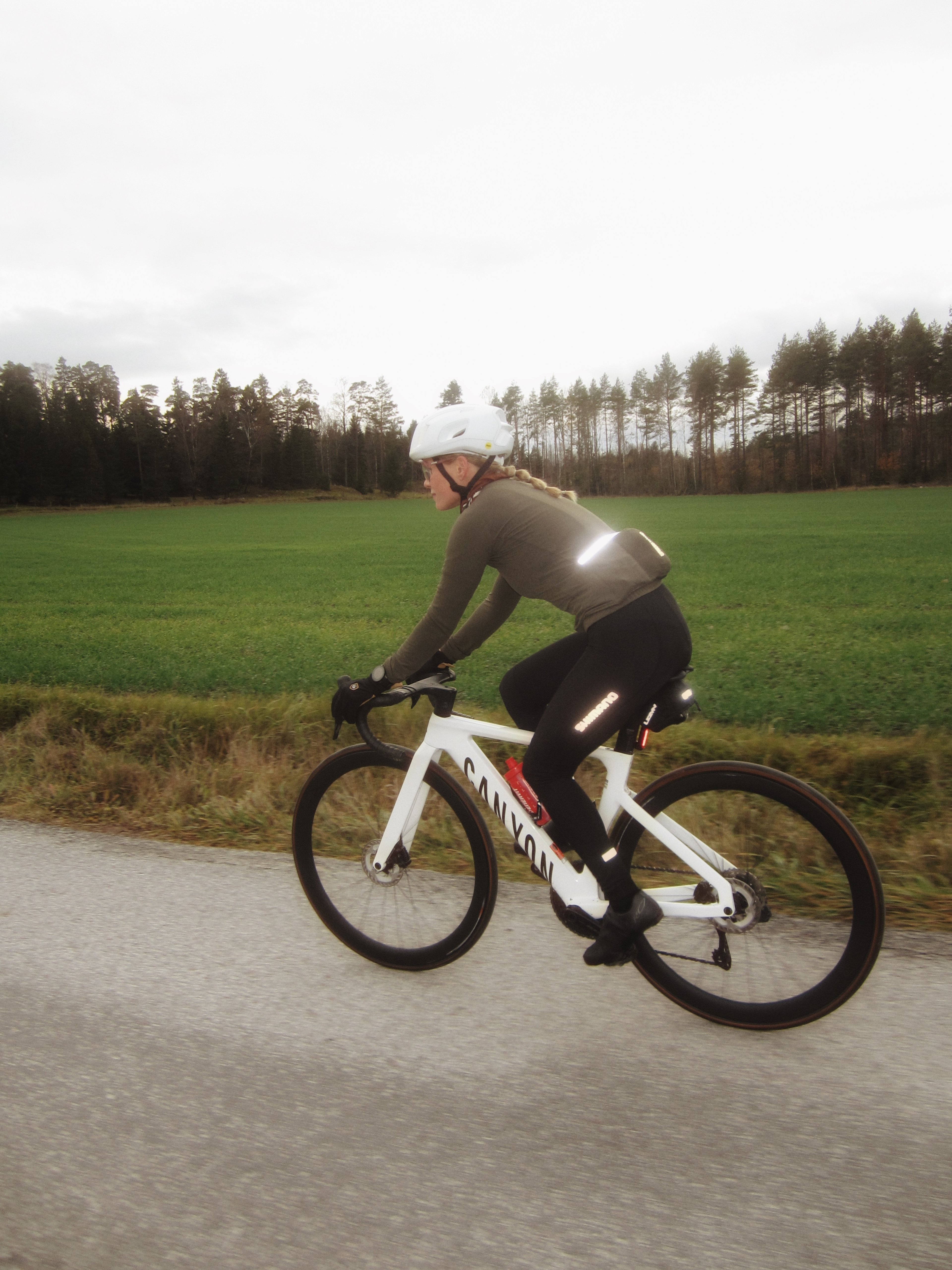 A woman in reflective cycling gear rides a white Canyon road bike on a paved road beside a green field and trees.