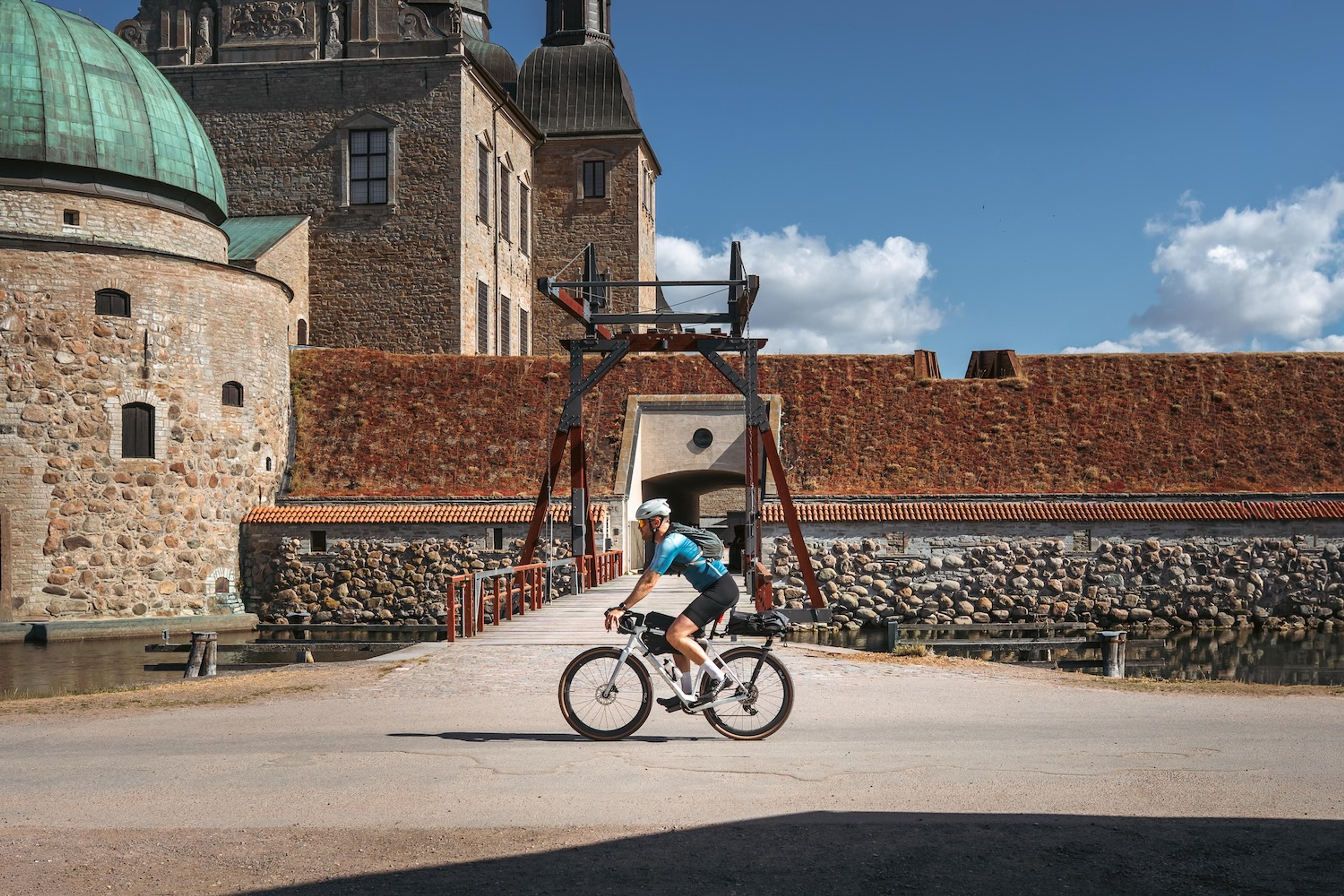 A person riding a bicycle crosses a drawbridge in front of a historic stone castle.