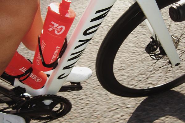 Close-up of a white Canyon bike with two orange Enervit water bottles, a cyclist's leg, and a front wheel on pavement.