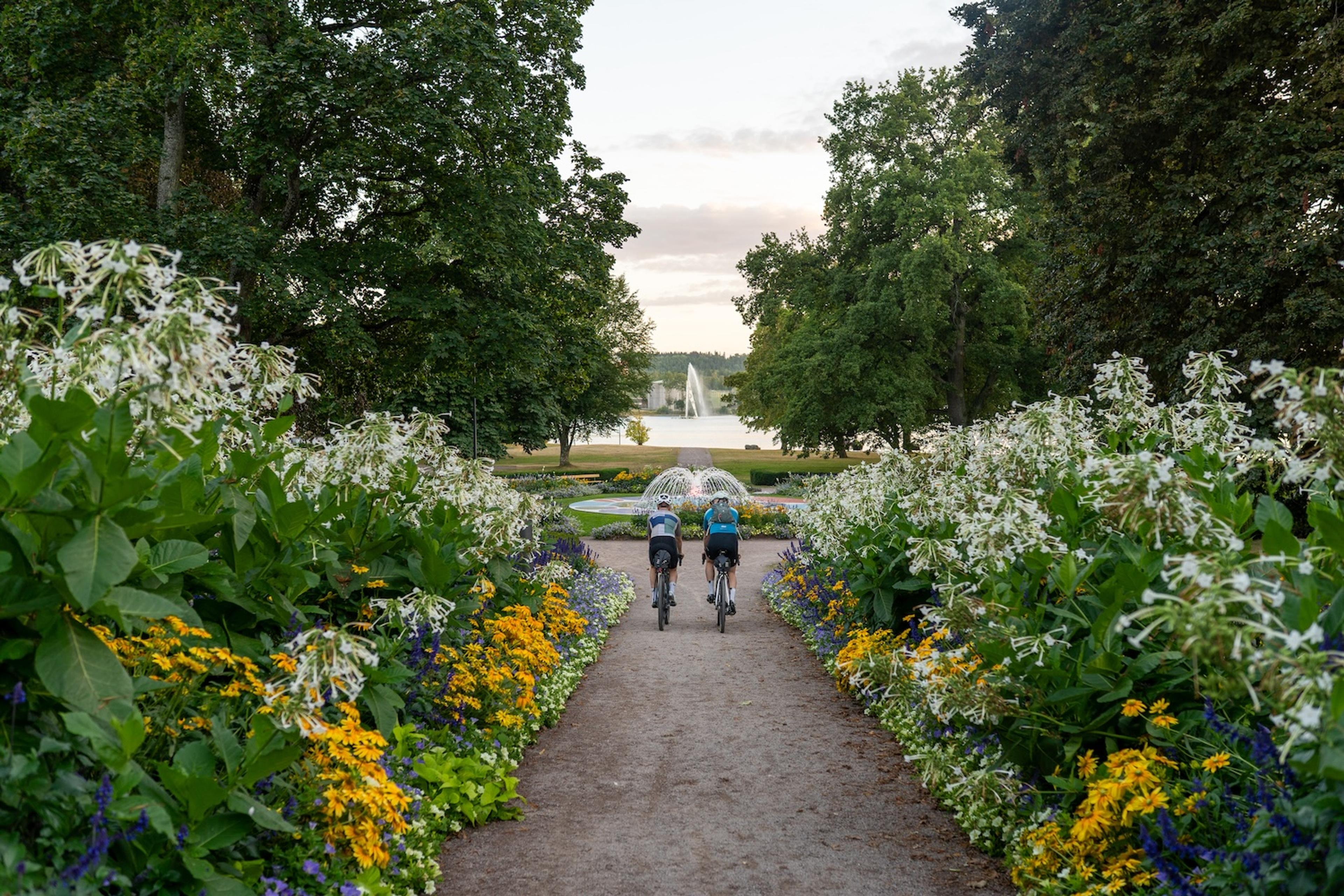 Two cyclists ride on a path lined with vibrant flowers towards fountains in a lake.