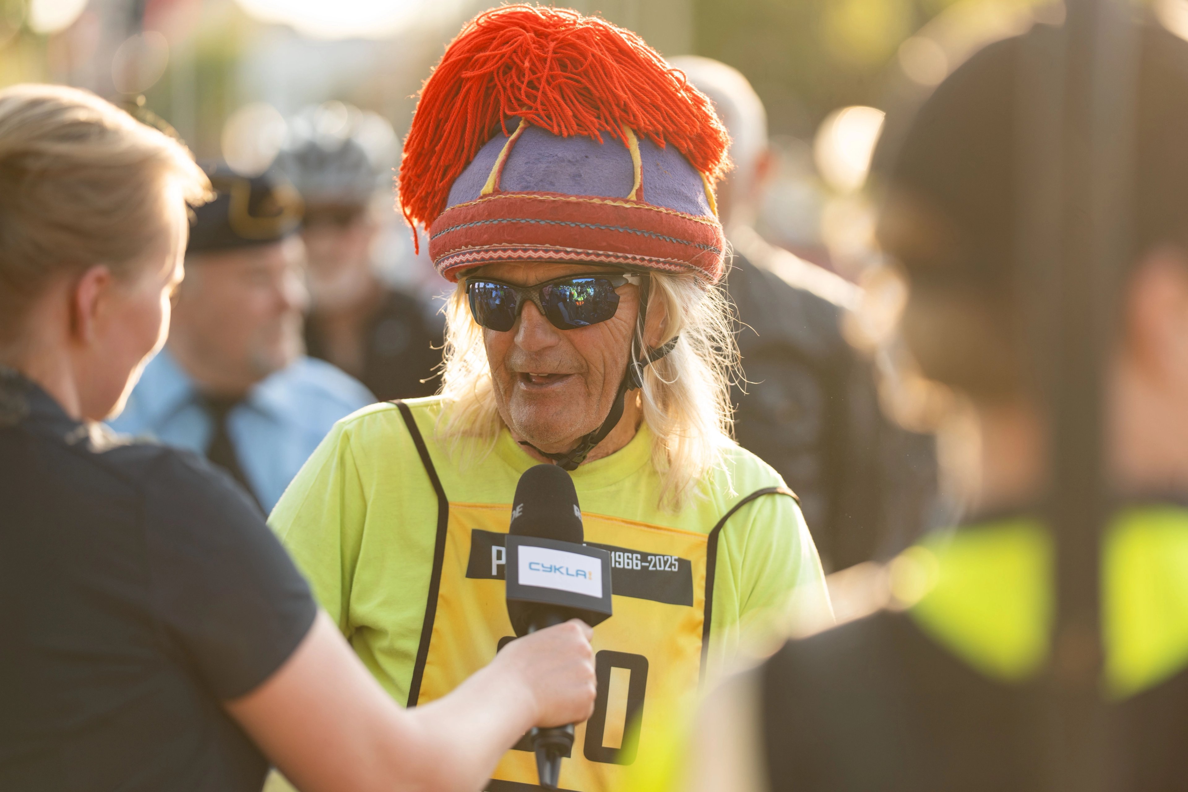 A man in a colorful helmet and yellow vest speaks into a microphone held by a woman.