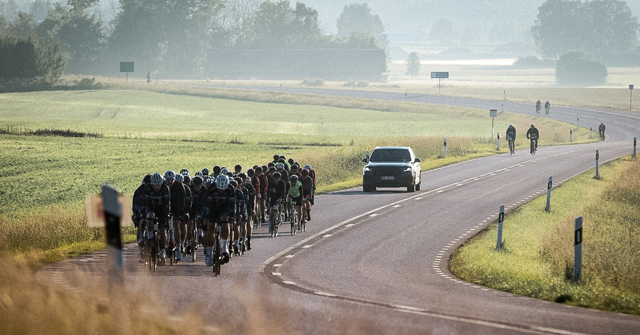 A large group of cyclists rides on a winding road with a support car in a hazy rural landscape.
