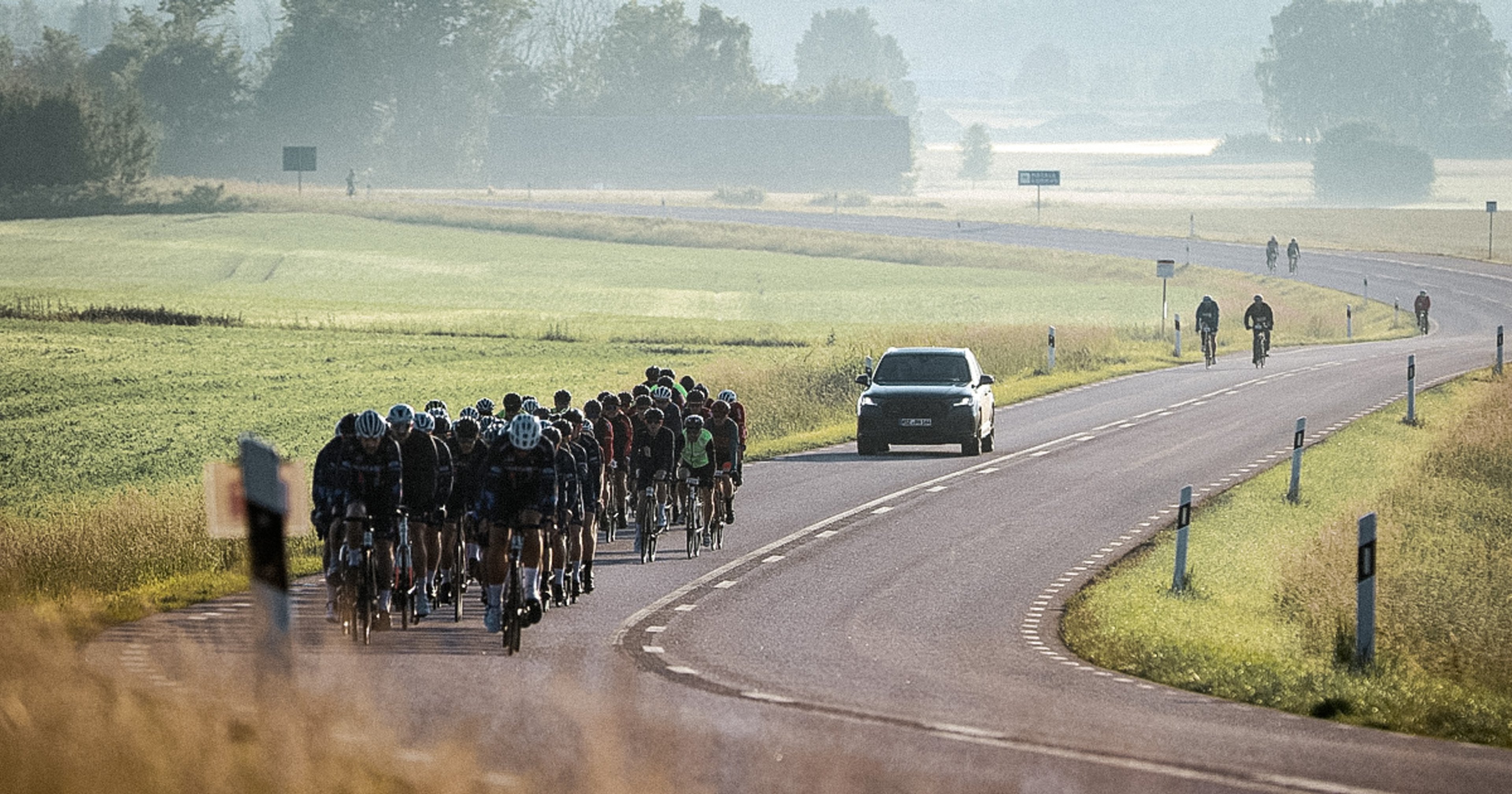 A large group of cyclists rides on a winding road with a support car in a hazy rural landscape.