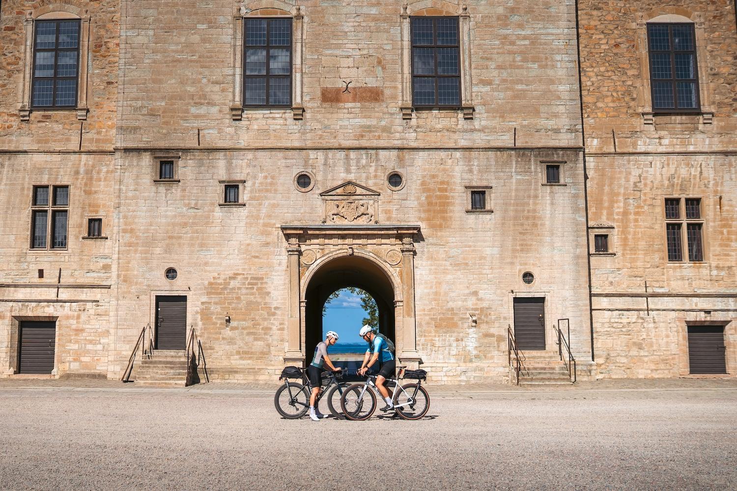 Two people with bicycles stand in front of a large stone building with an archway revealing the ocean.