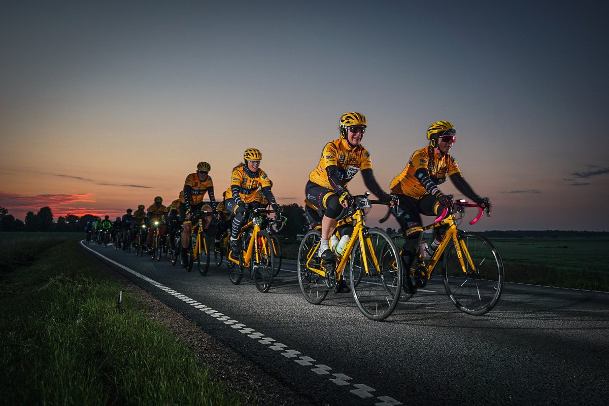Five happy cyclists ride on a road bordered by trees, with one person waving and another making a funny face.