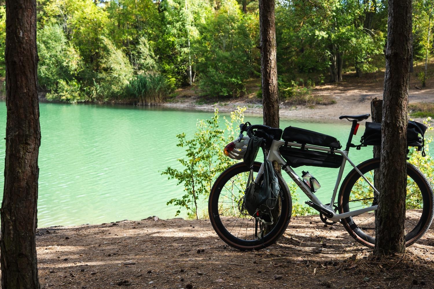 A bikepacking bike stands next to a turquoise lake in a forest.