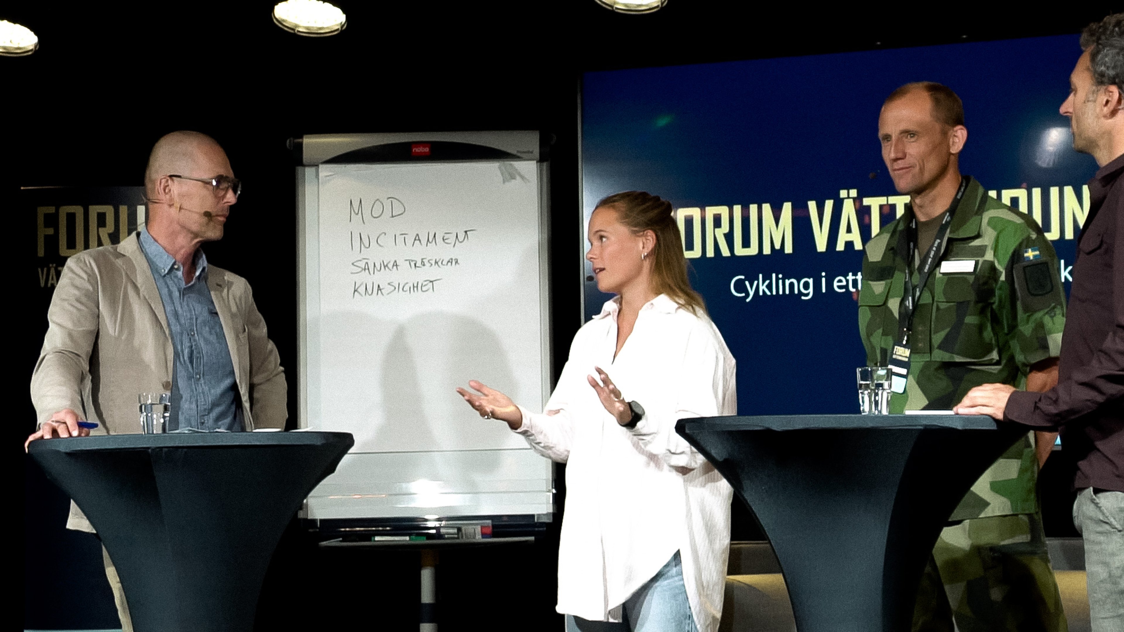 Four people on a stage at a panel discussion, with a whiteboard and a banner in the background.