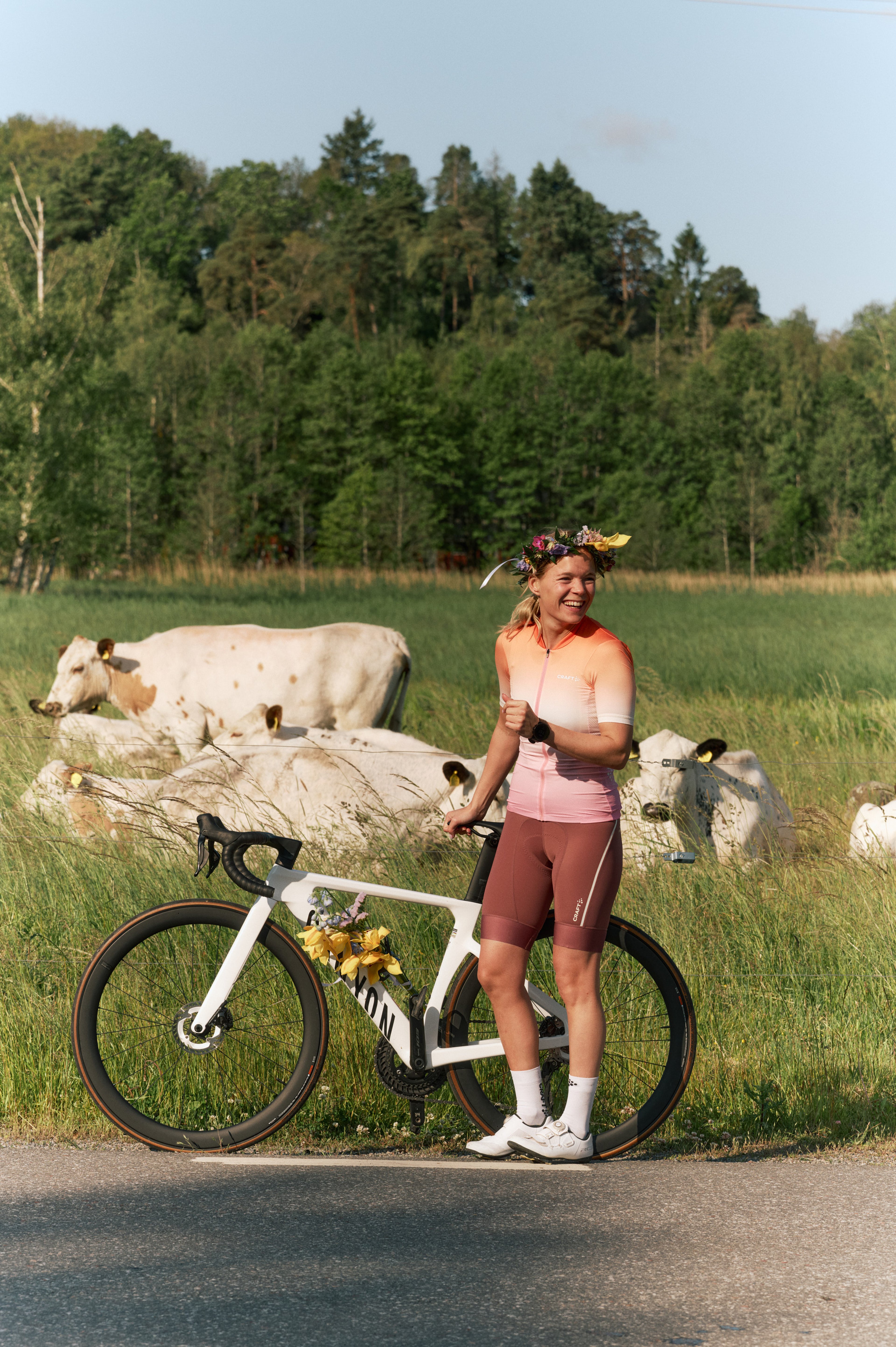 A smiling woman in cycling gear and a flower crown stands next to her white bicycle decorated with flowers, giving a thumbs up in a field with cows.