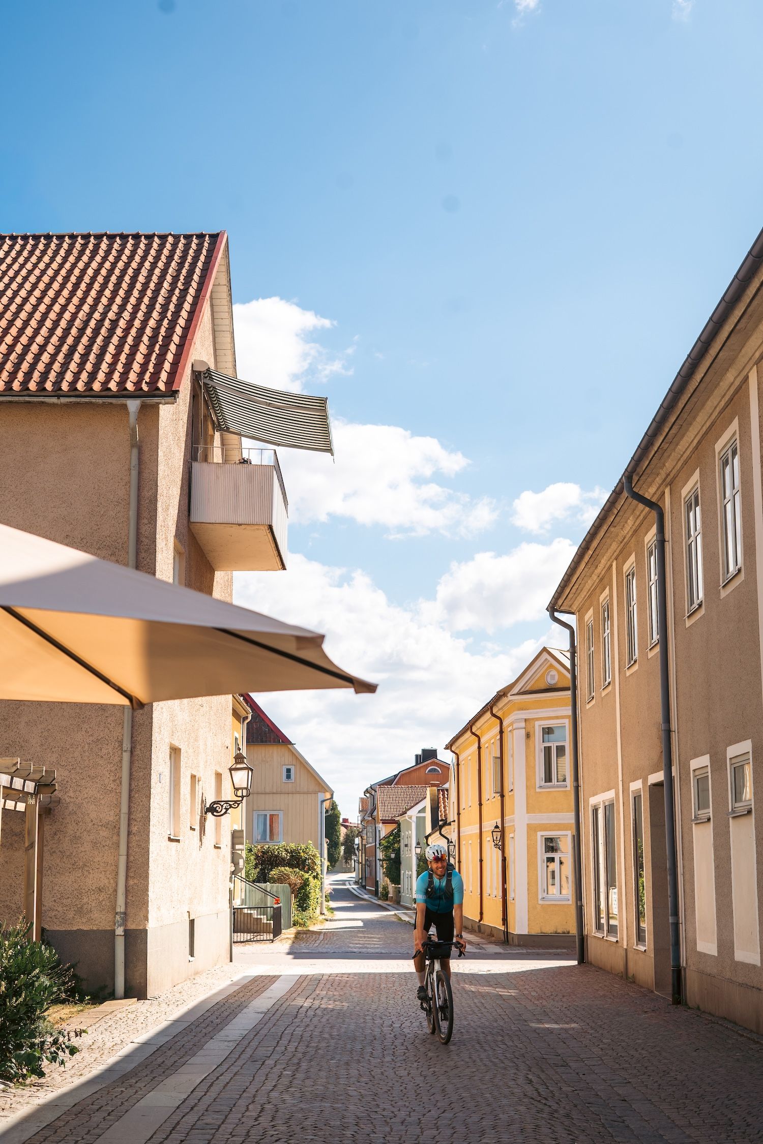 A person cycles down a sunny, cobbled street lined with colorful historic buildings.