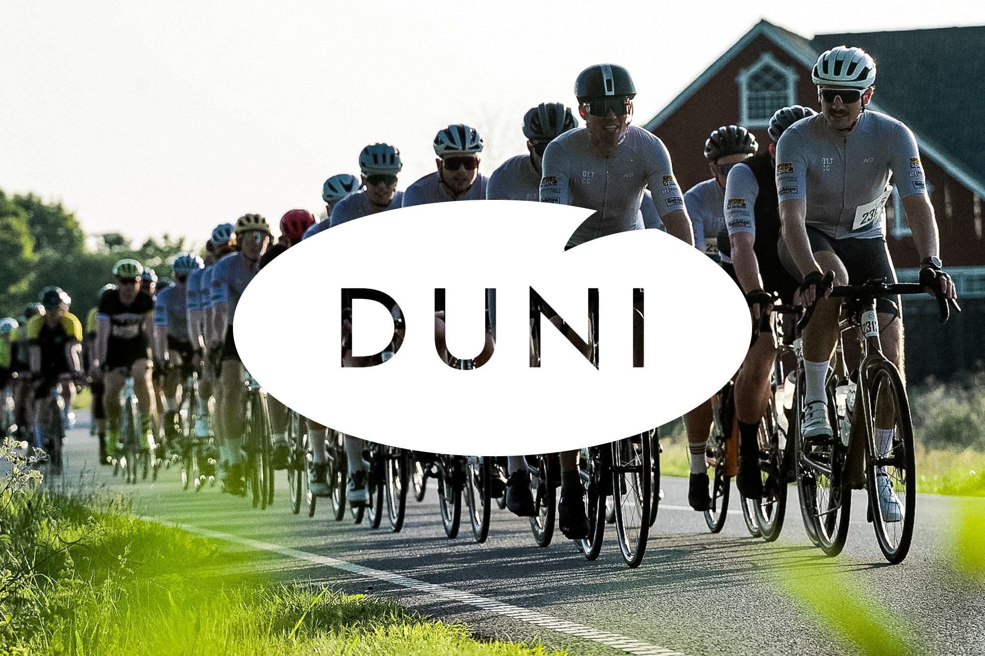 Cyclists ride on a rural road with a red barn in the background, featuring a white speech bubble overlay with 'DUNI'.