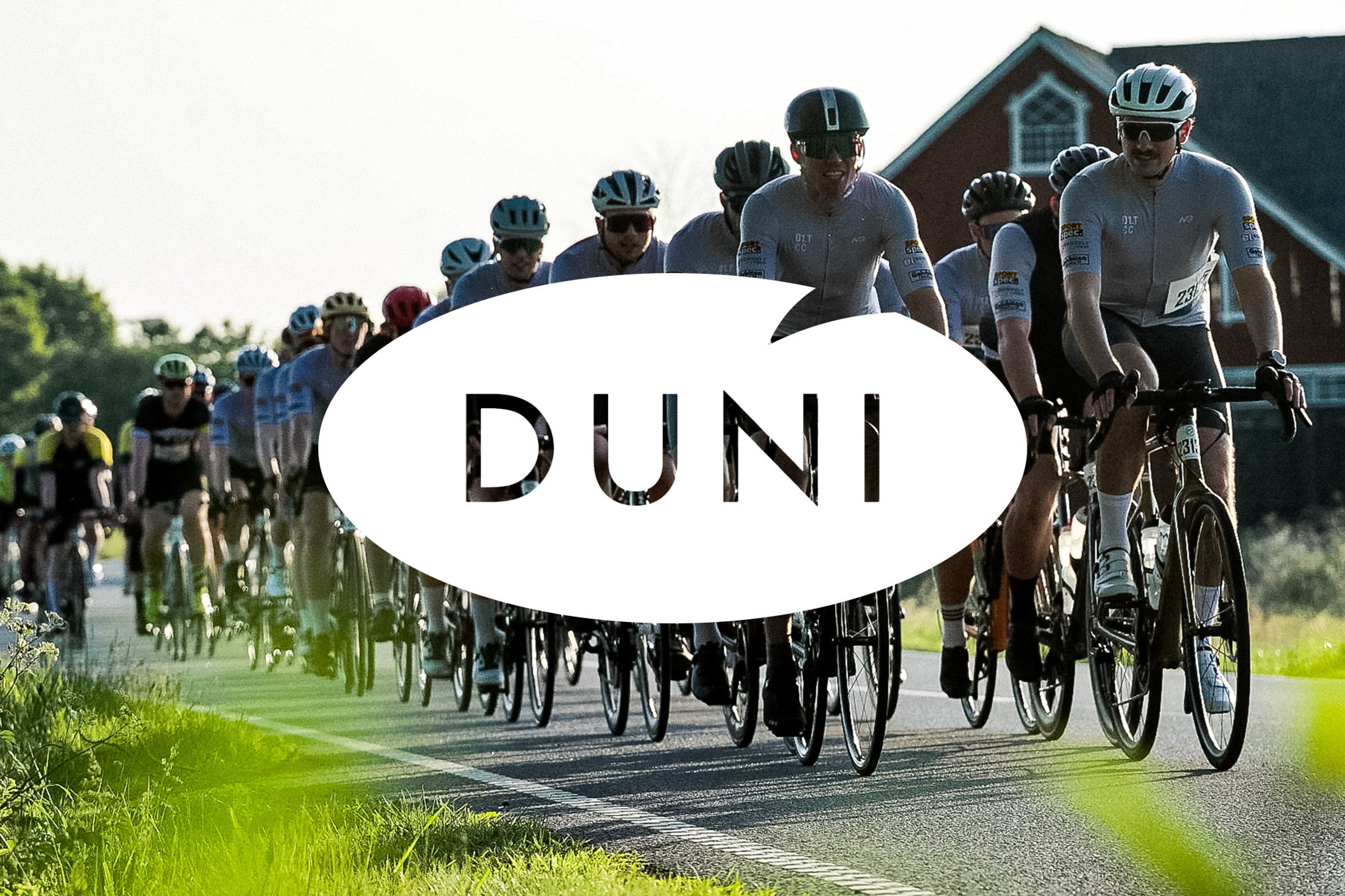 Cyclists ride on a rural road with a red barn in the background, featuring a white speech bubble overlay with 'DUNI'.