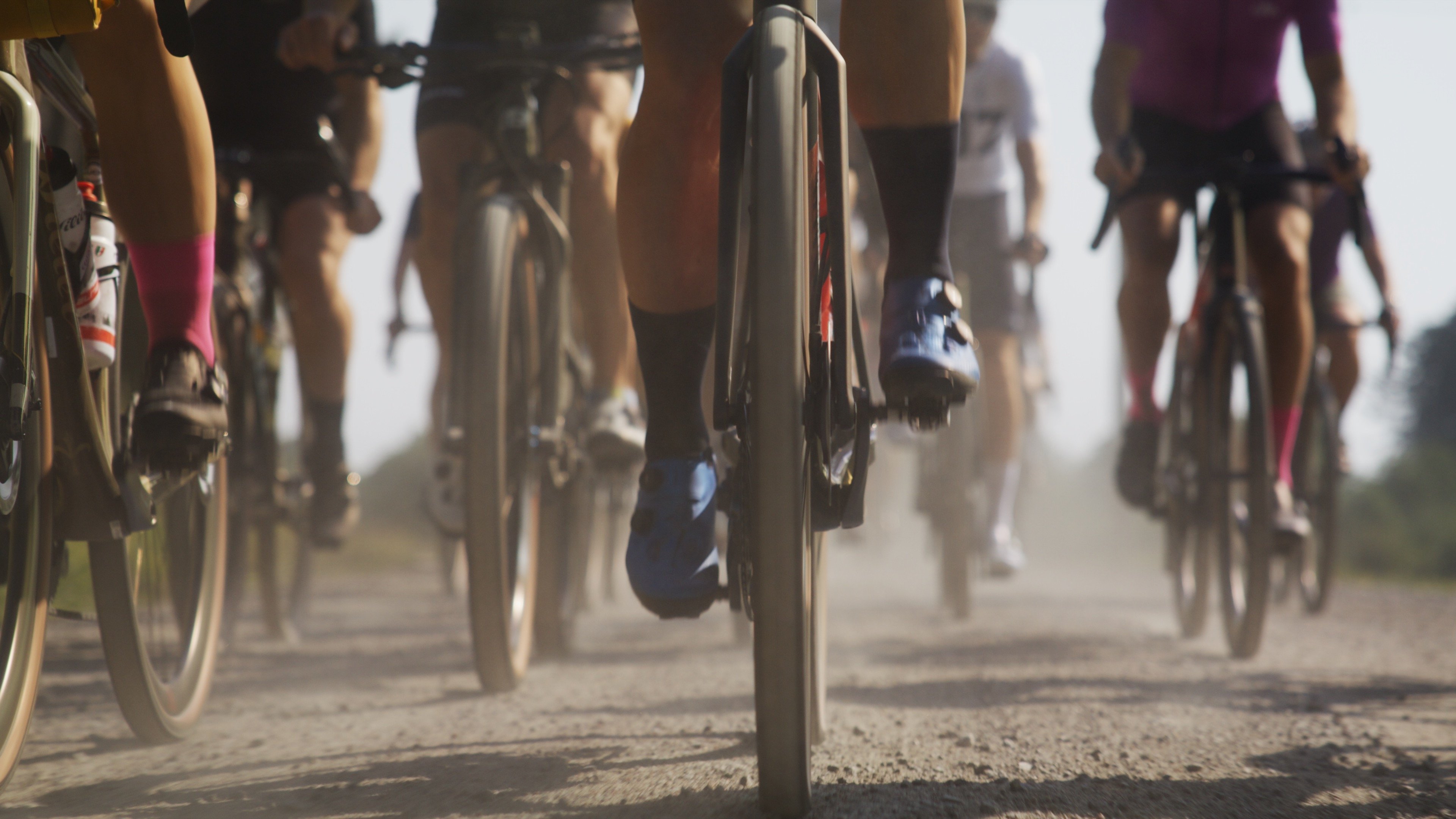 Cyclists' legs and wheels kicking up dust on a gravel road.
