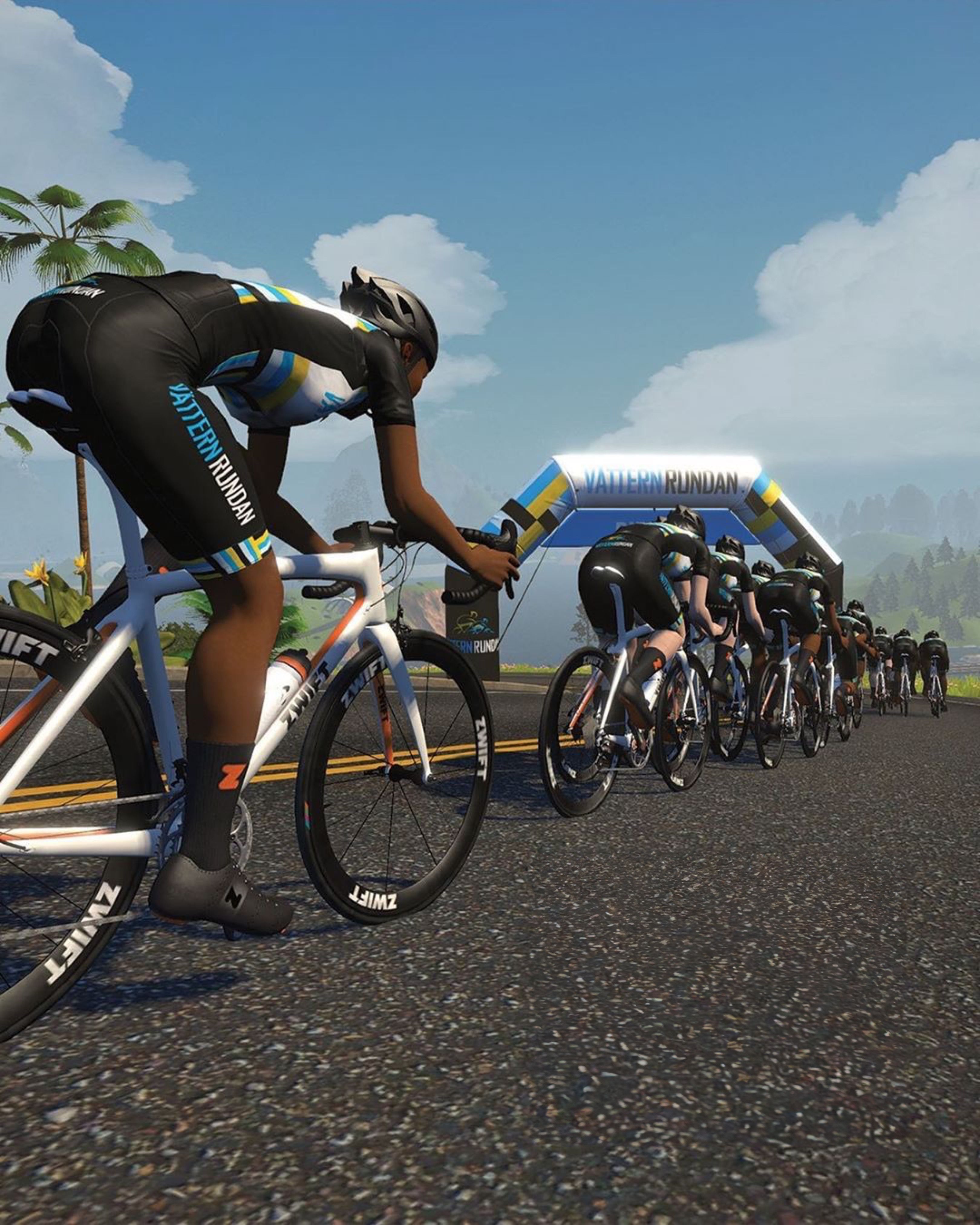A virtual cyclist in a black, blue, and yellow "Pattern Rundan" kit leads a pack of riders on a road towards a "Vätternrundan" archway under a blue sky.