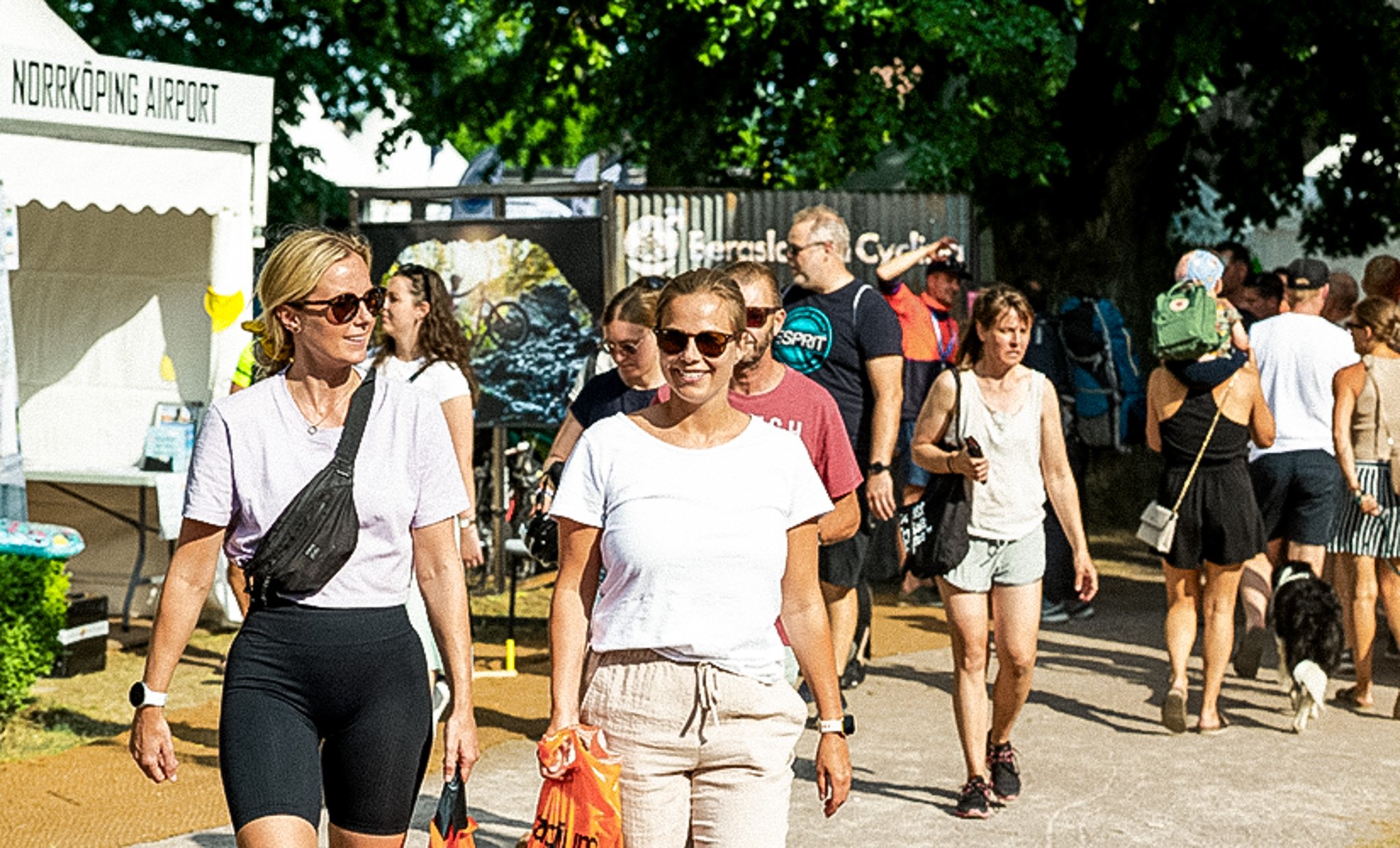 Two smiling women walk among a crowd at a sunny outdoor event.
