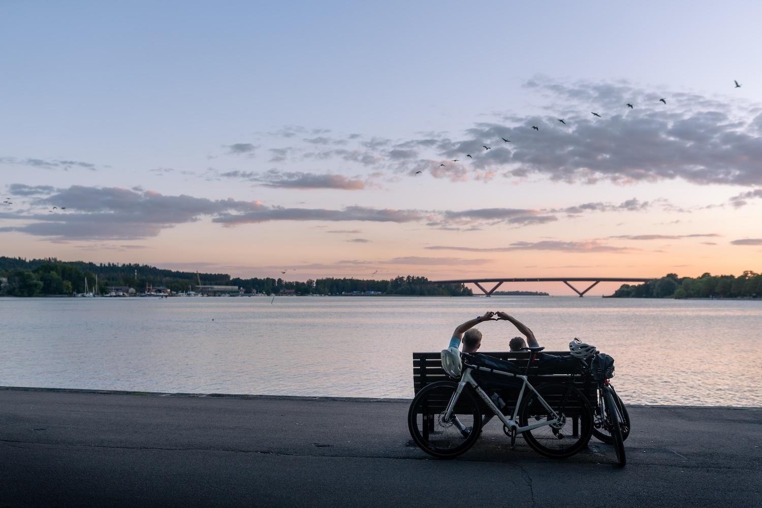 A couple makes a heart shape with their hands on a bench next to bikes, overlooking a lake with a distant bridge and flying birds at sunset.