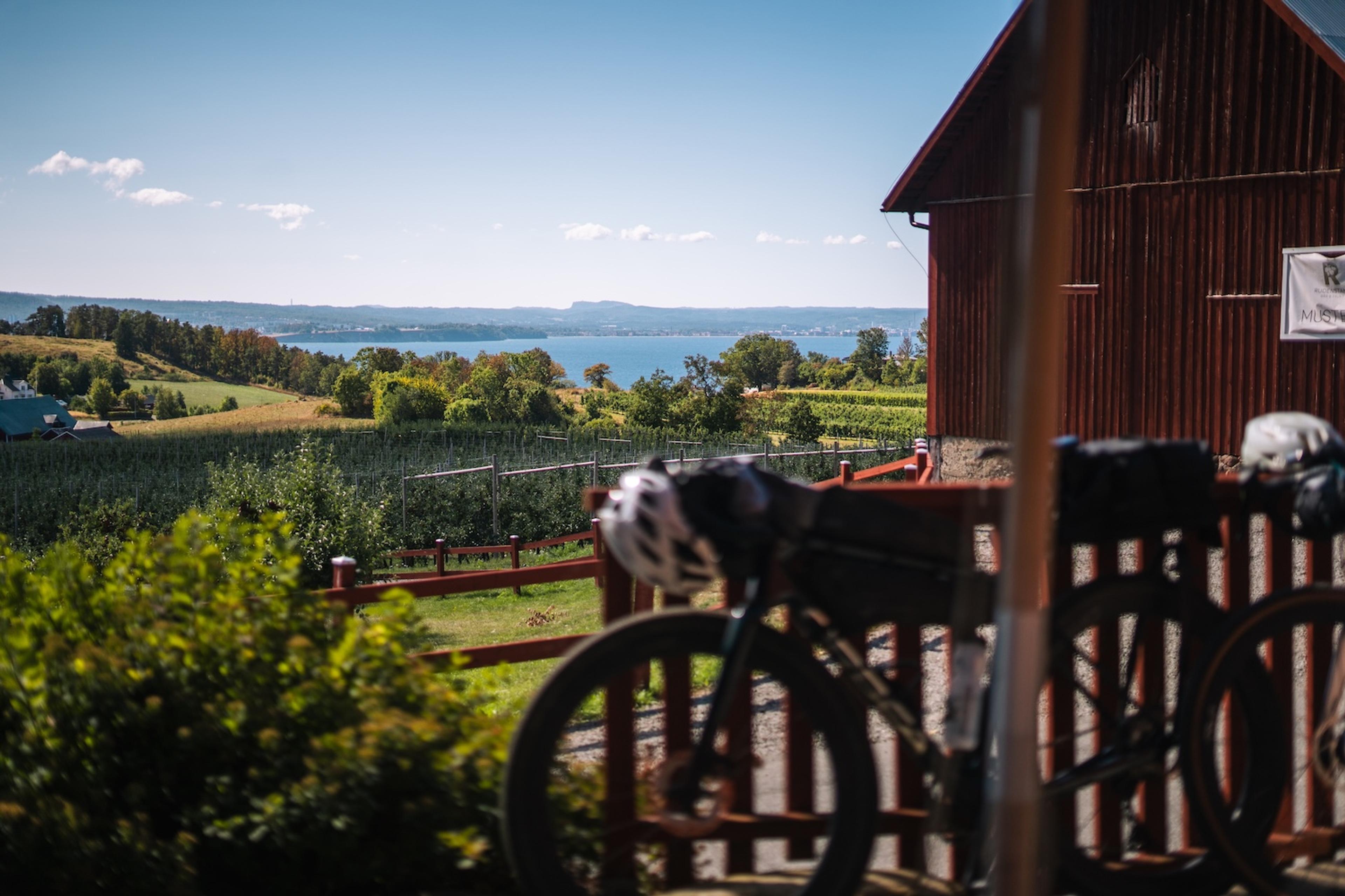 Two touring bicycles parked at a fence overlooking a lake, orchards, and a red barn under a blue sky.