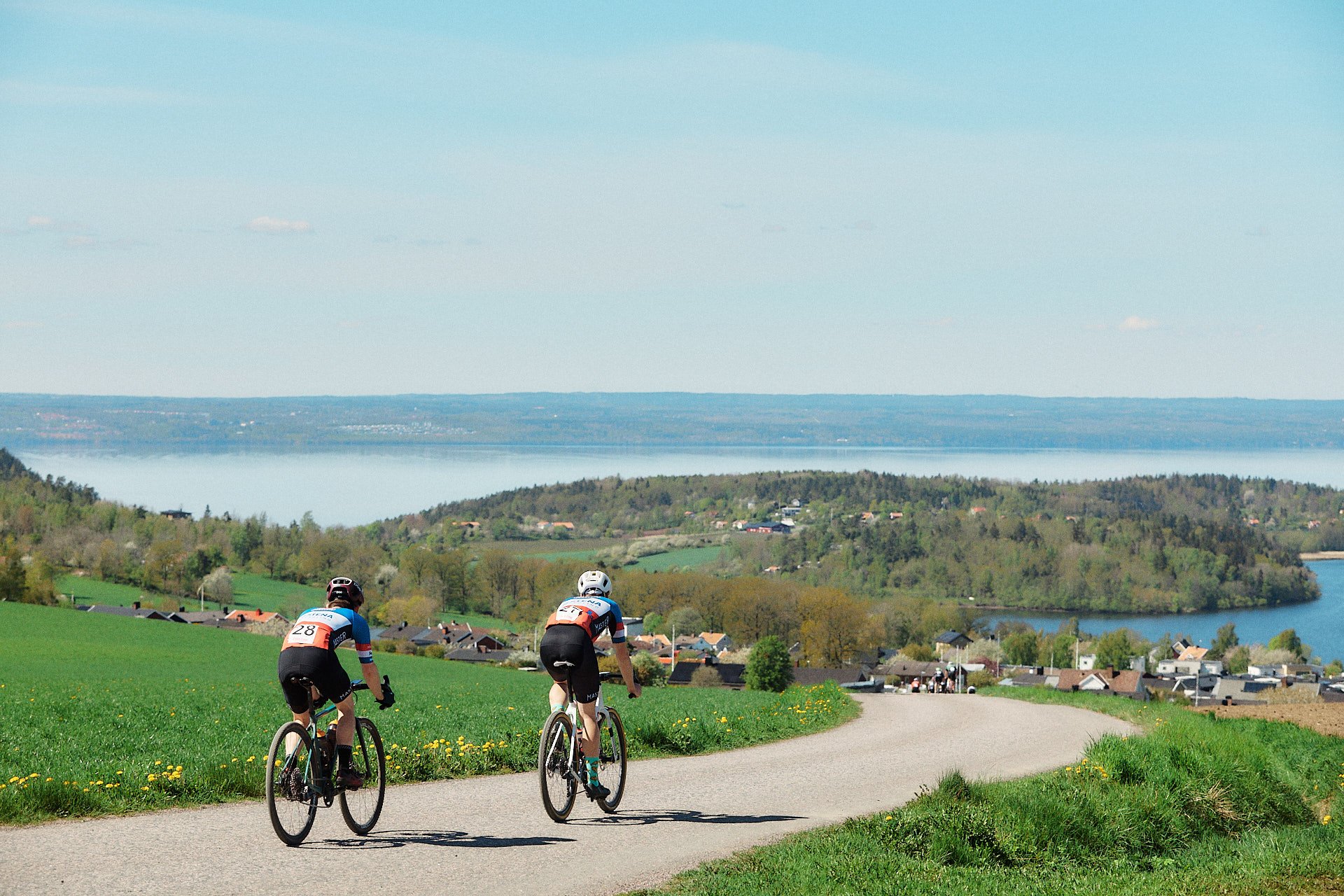 Two cyclists ride on a road with a scenic view of a large lake and green hills.