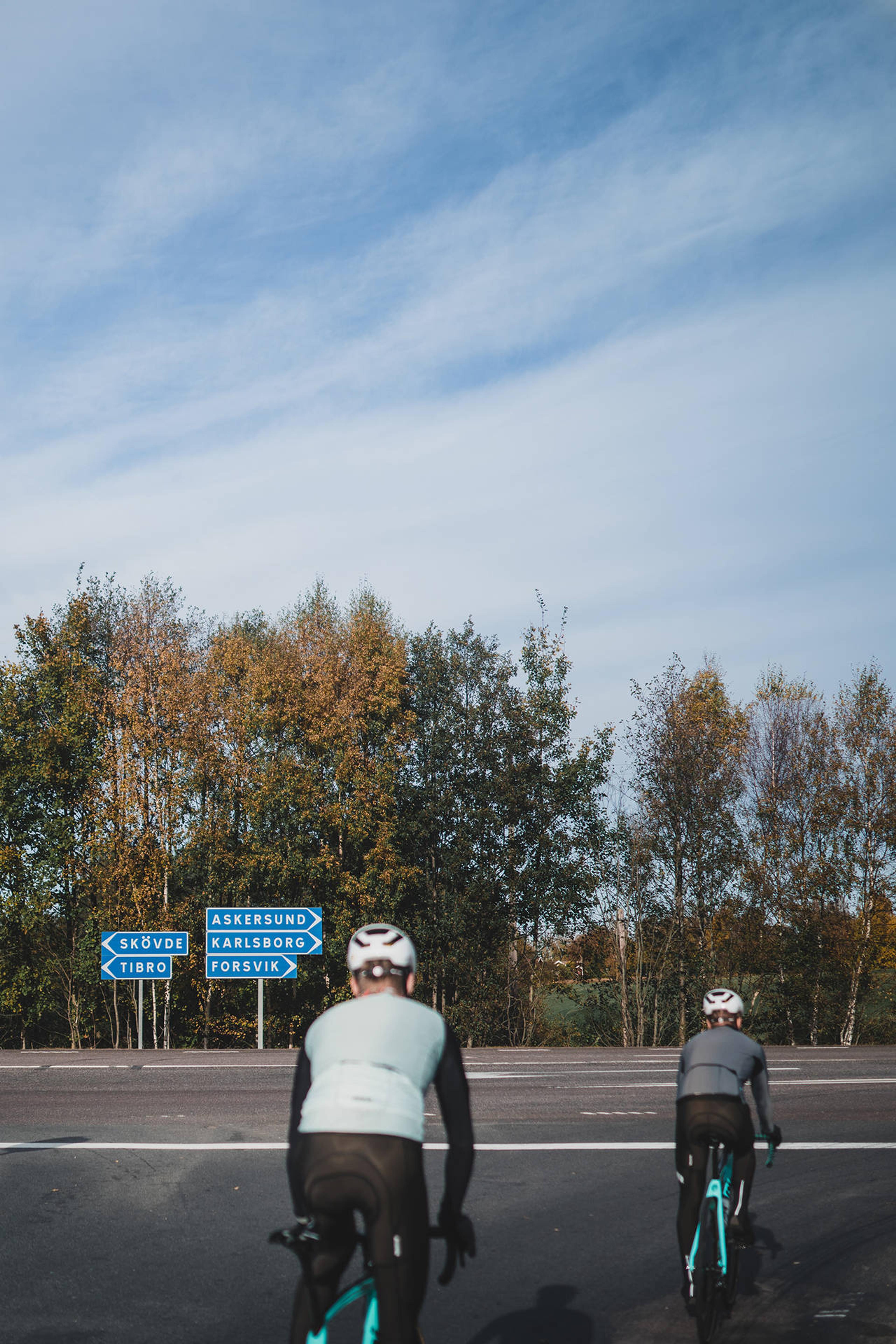 Two cyclists ride away from the viewer on a road, approaching blue directional signs for various towns. Trees with fall foliage are in the background under a cloudy blue sky.