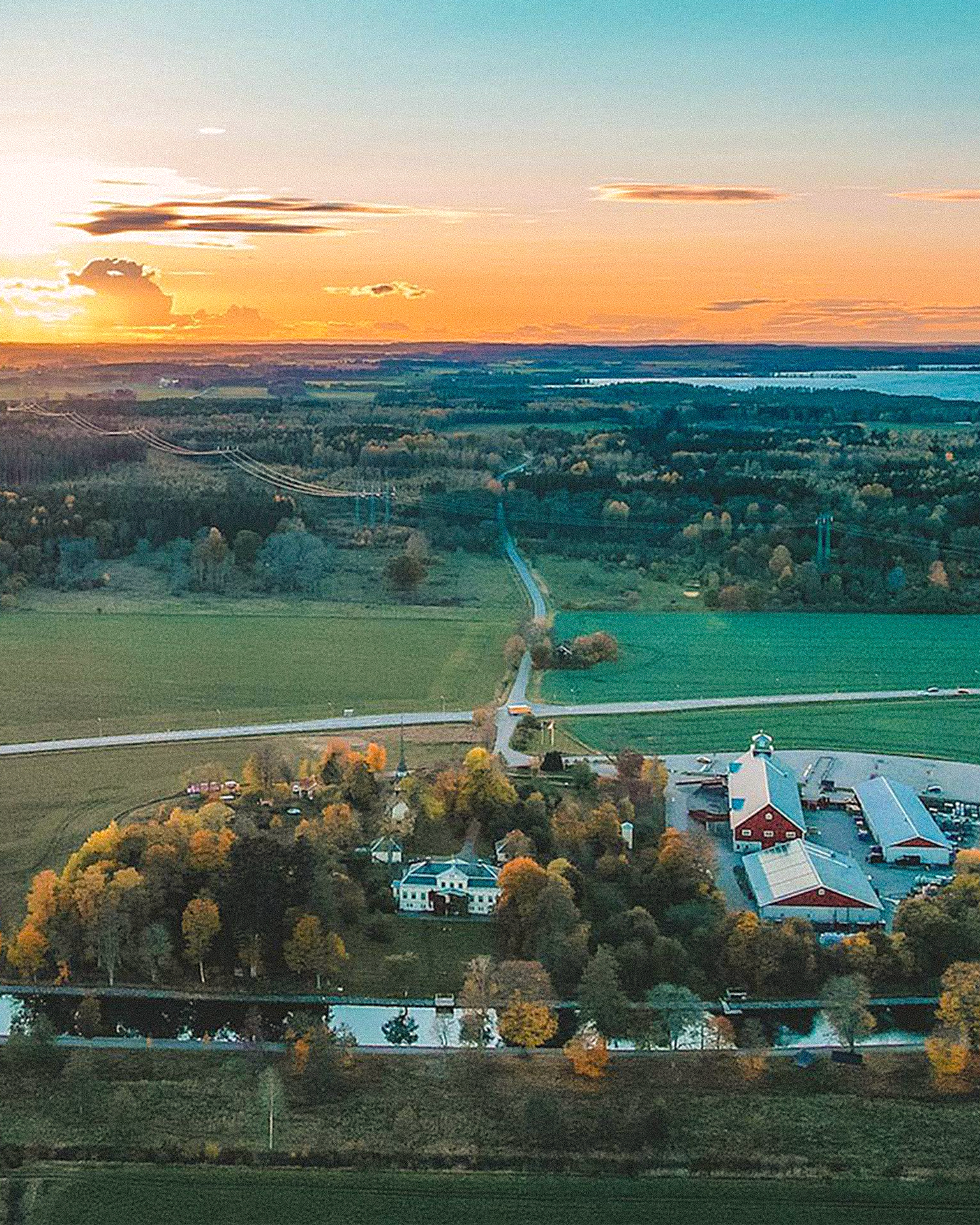 Aerial view of a rural landscape at sunset, showing green fields, forests, a canal, a white mansion, red-roofed buildings, and a distant lake under an orange and blue sky.