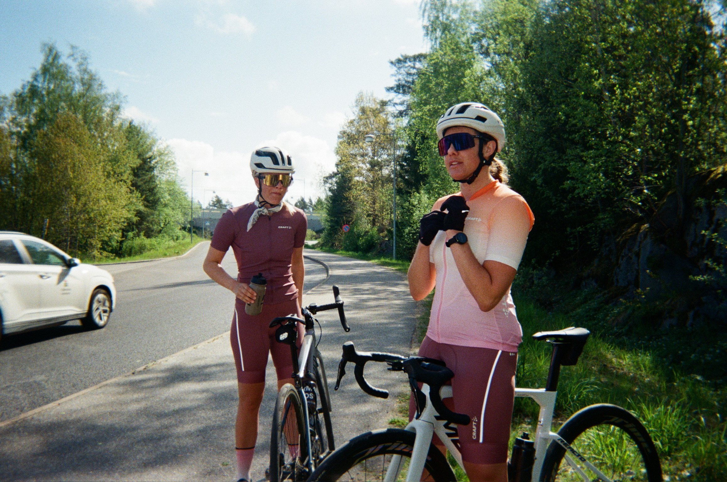 Two female cyclists in helmets and colorful kits stand with their bikes on a sunny road.