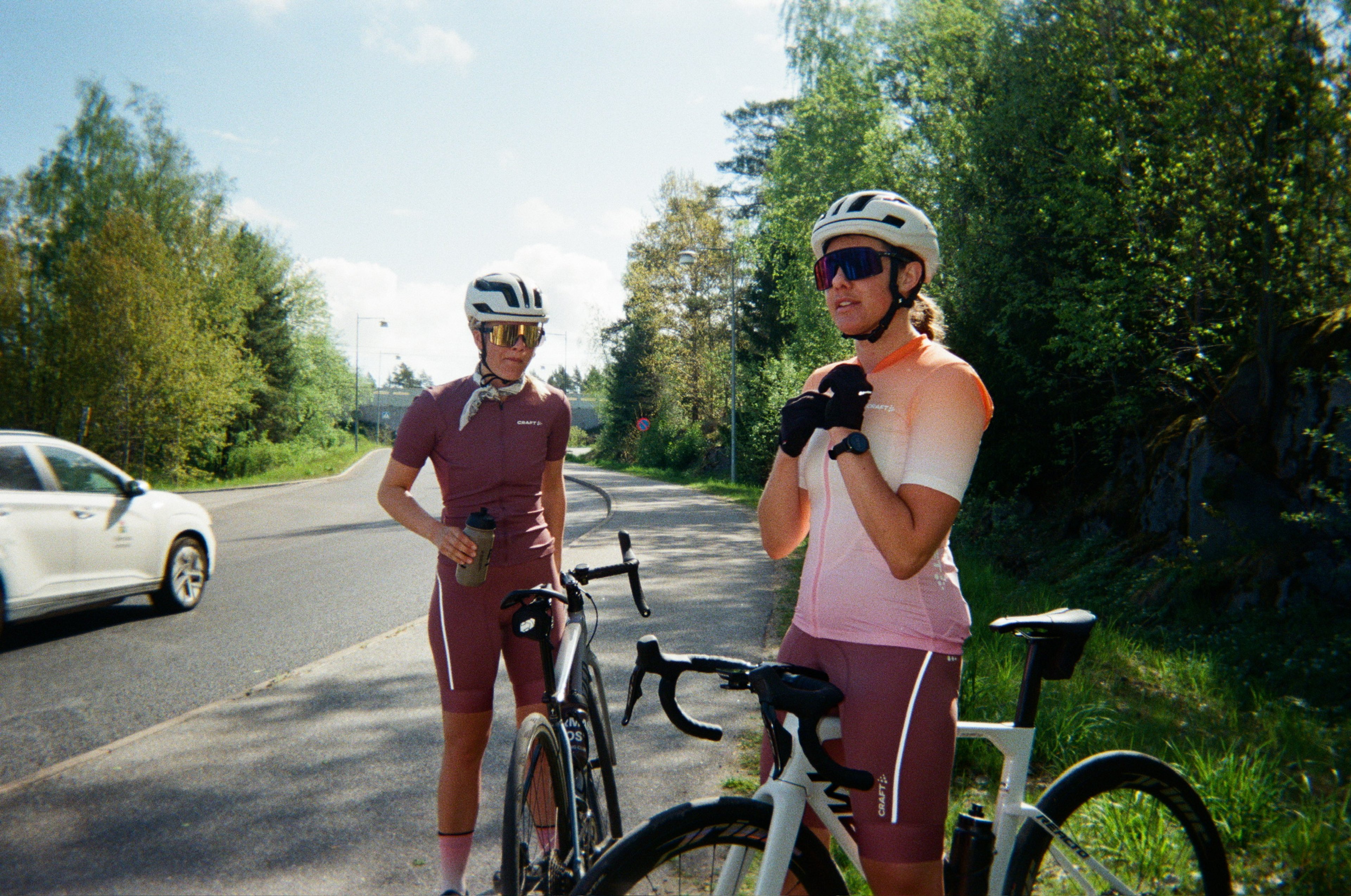 Two female cyclists in helmets and colorful kits stand with their bikes on a sunny road.